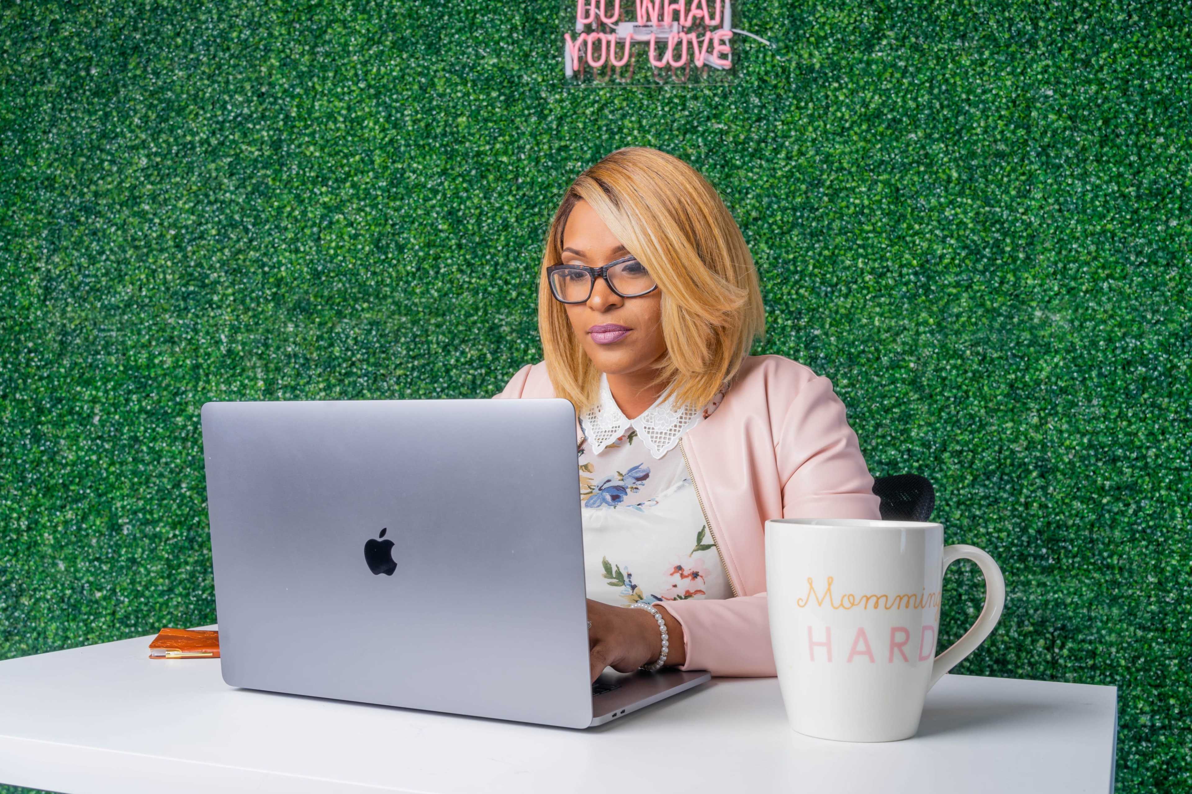 A woman with blonde hair works on a laptop at a white table in front of a green wall, with a large coffee mug nearby that reads "Mommin' Hard."