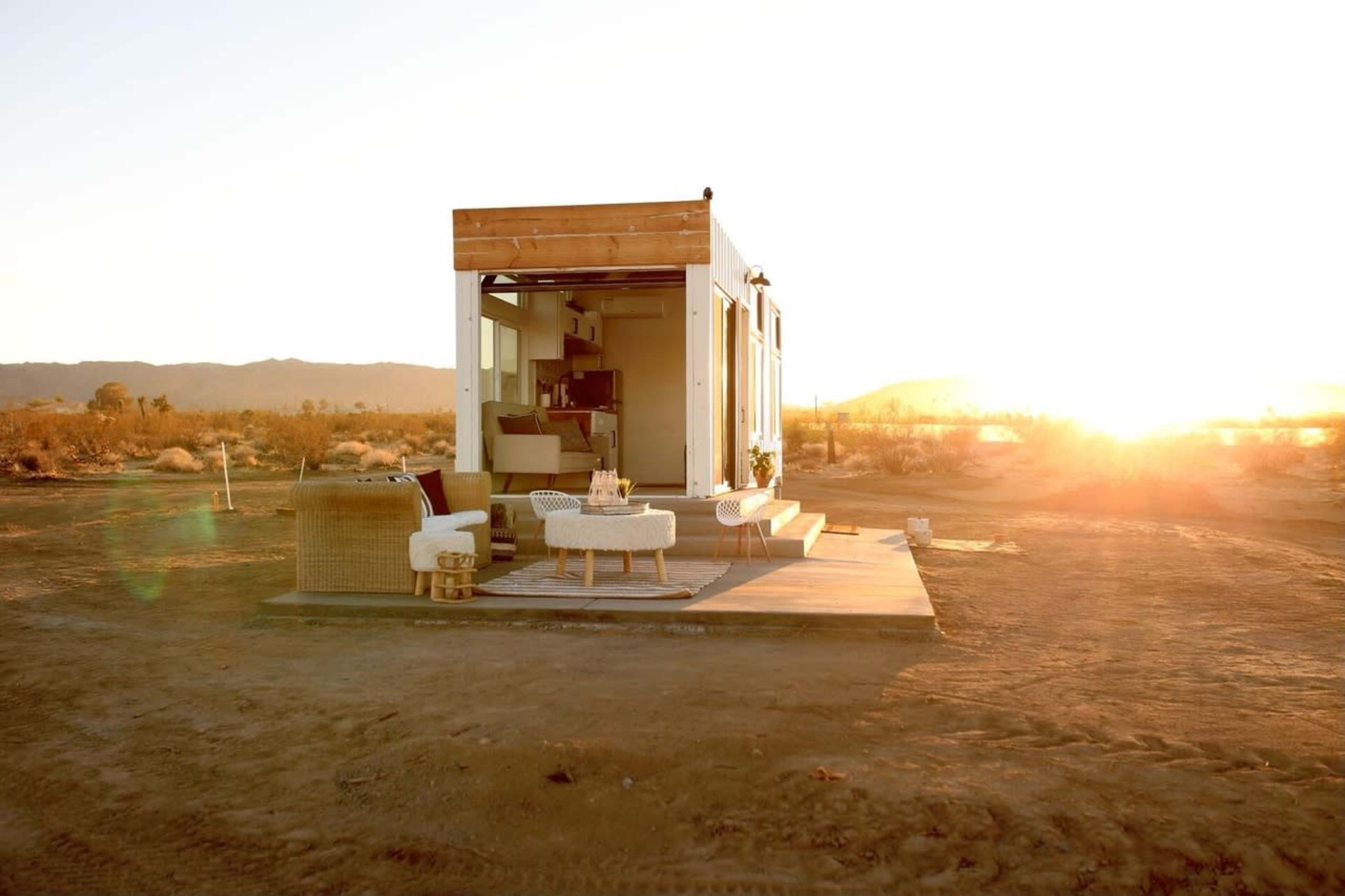 A modern tiny house sits on a flat area of desert land at sunset, with its doors open to reveal a cozy interior.