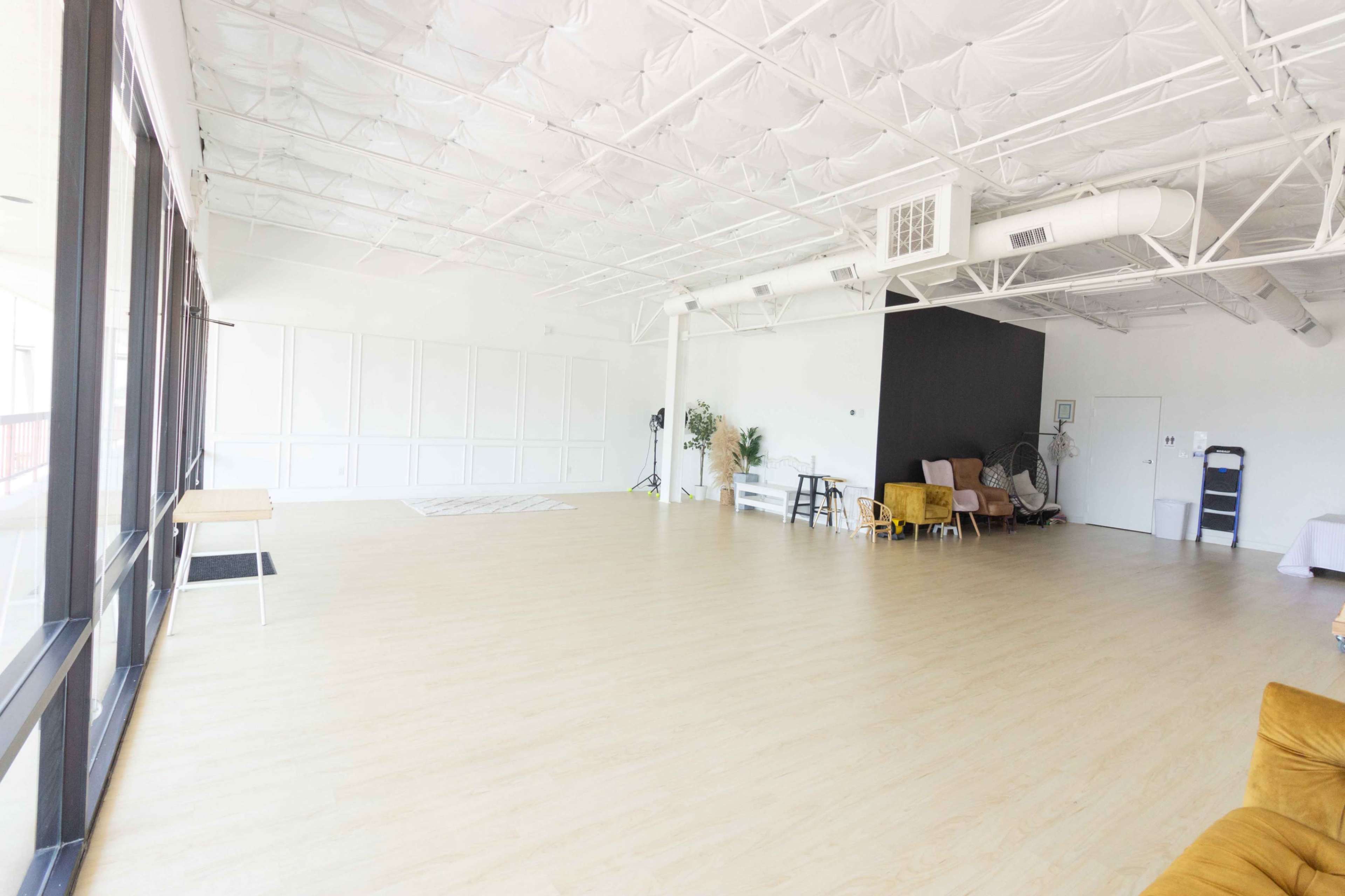 A spacious, empty room with a wood floor, a black accent wall, and minimal furnishings, including a small table and a few chairs.