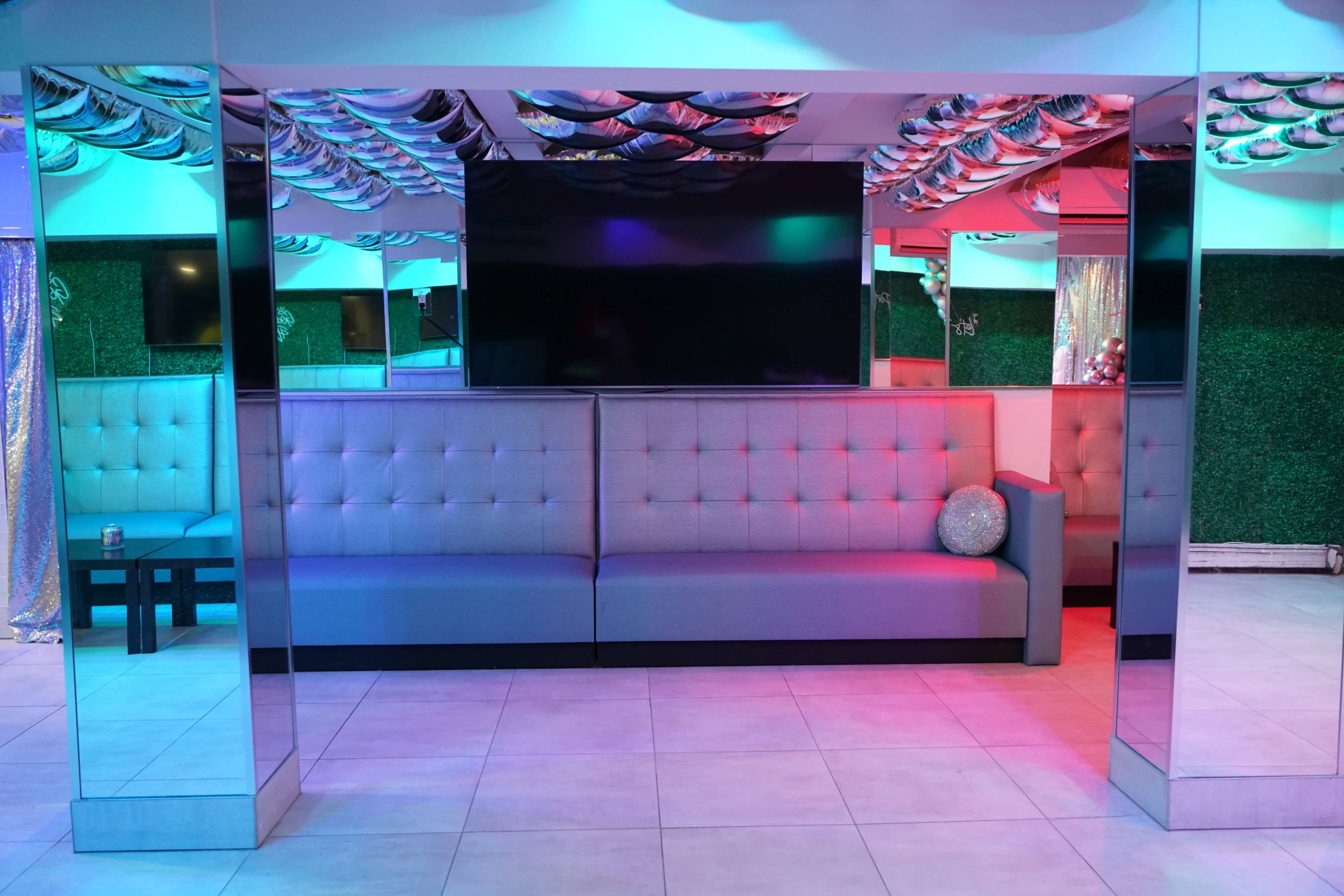 A modern lounge area with a gray sectional couch, a large television on the wall, and colorful lighting above.