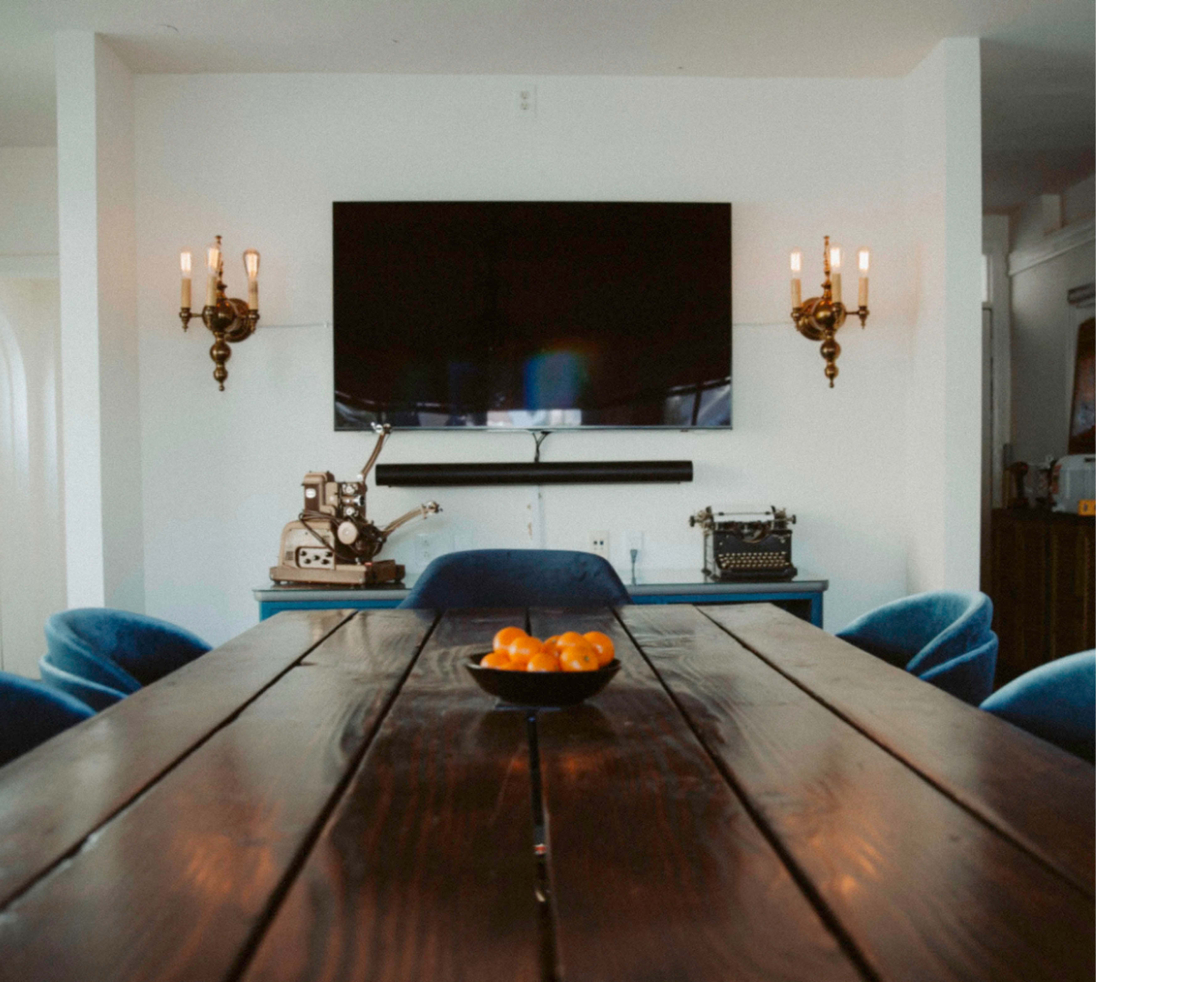 A wooden dining table is centered in a room with a wall-mounted television above, flanked by wall sconces, and a bowl of oranges sits in the middle.