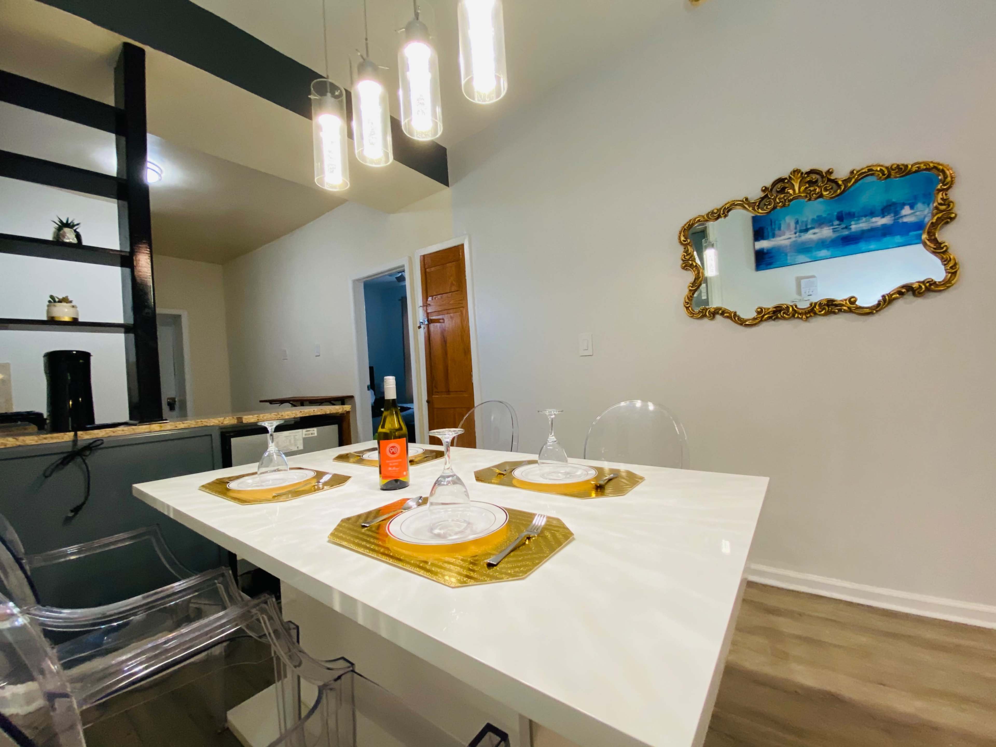 A modern dining area features a white table set with plates and glasses, accompanied by a bottle of wine, with pendant lights hanging above and a mirror on the wall.