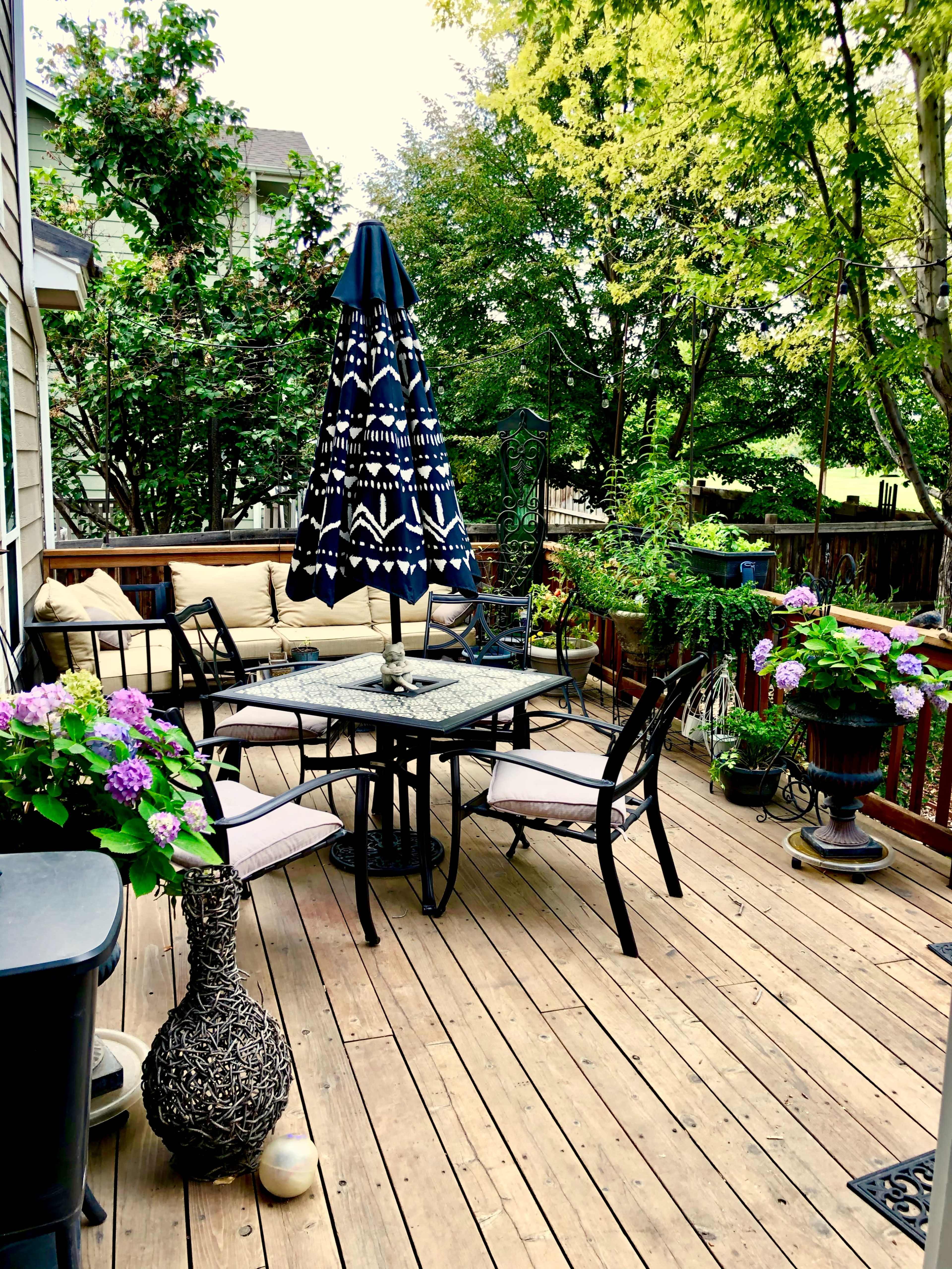 A patio setting features a round table with four chairs, an umbrella, and various potted plants in a wooden deck area.