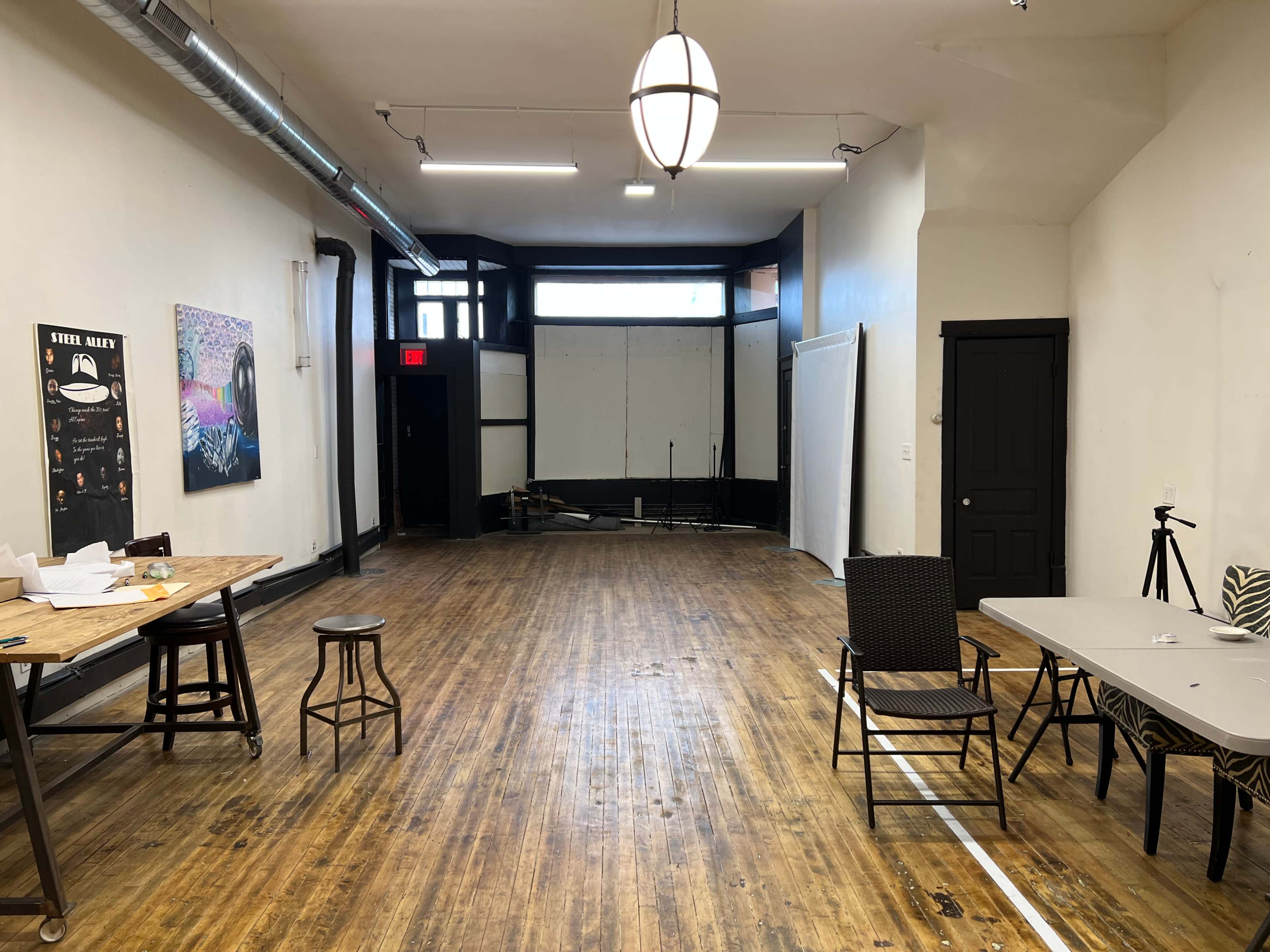 An empty studio space with wooden floors, a large window at the front, and various pieces of furniture scattered around, including a table and chairs.