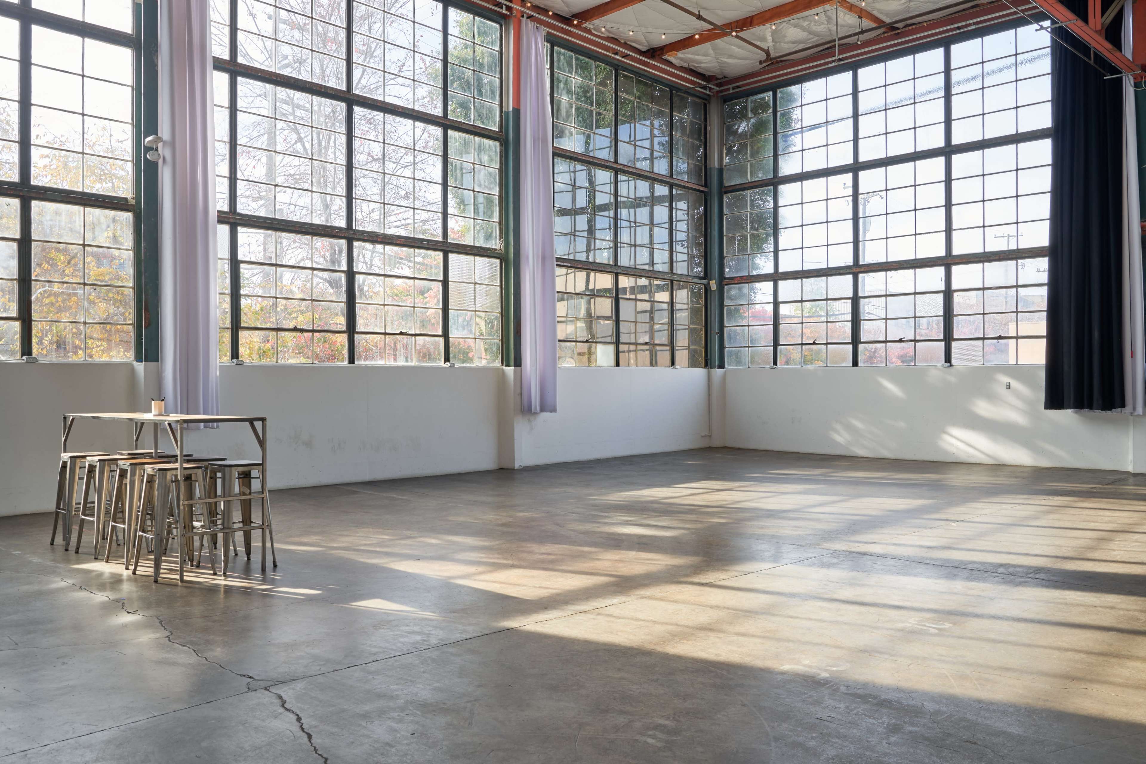 An empty industrial space with large windows and a set of metal stools arranged on a polished concrete floor.