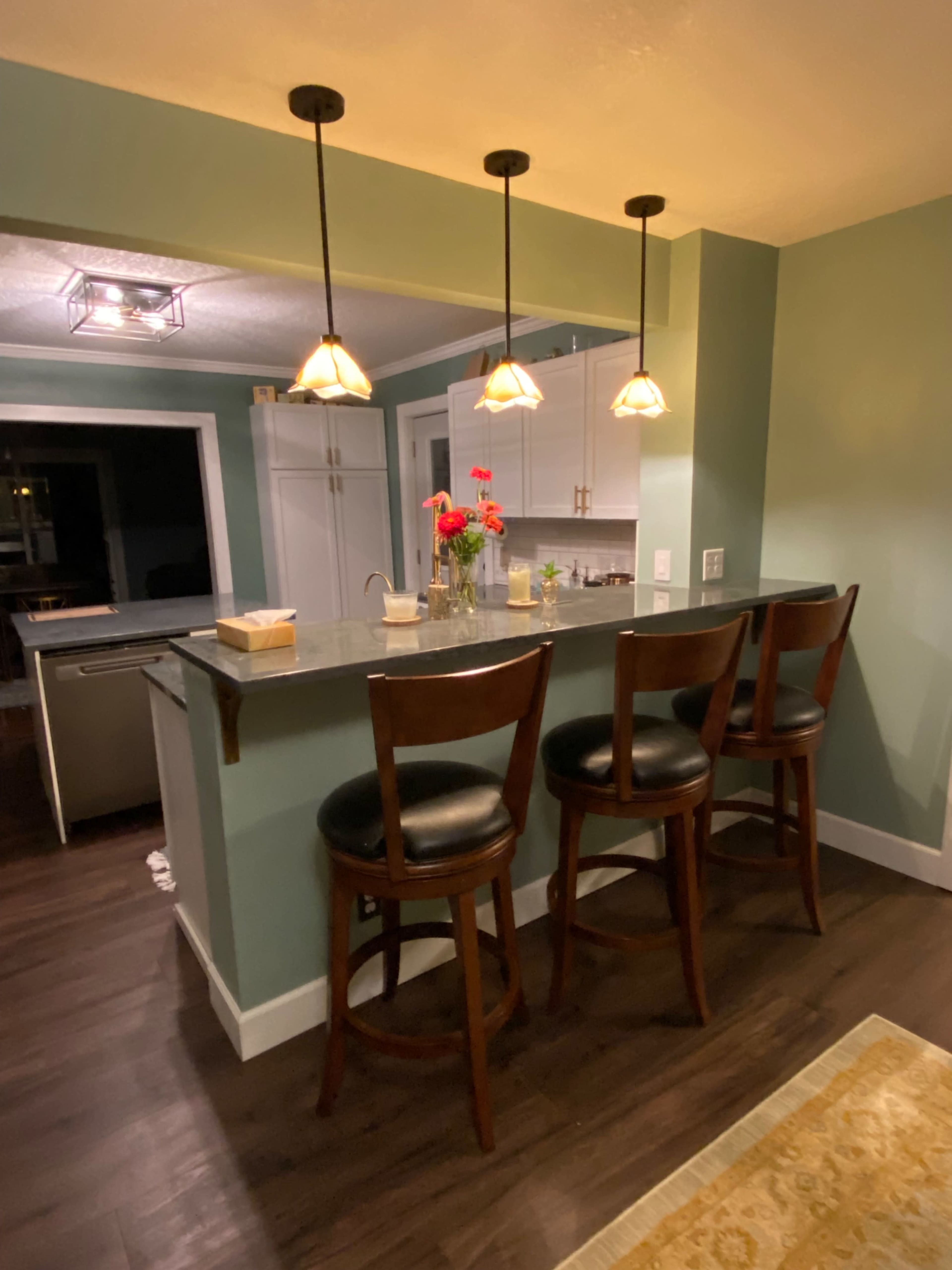 The image shows a kitchen bar area with three wooden stools, pendant lighting above, and a countertop decorated with a vase of flowers.