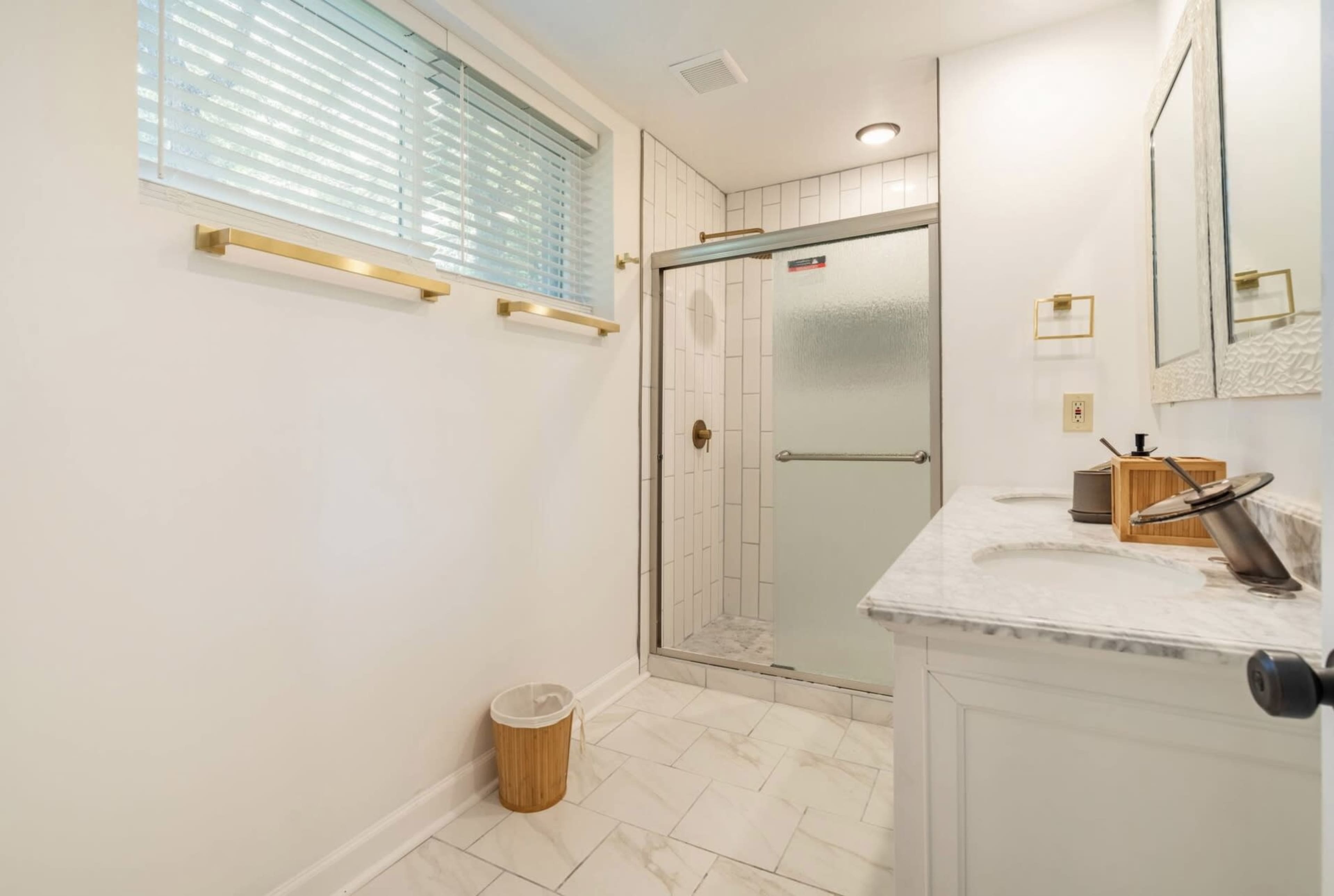 The bathroom features a glass shower, a marble countertop, and natural light coming through a window with blinds.