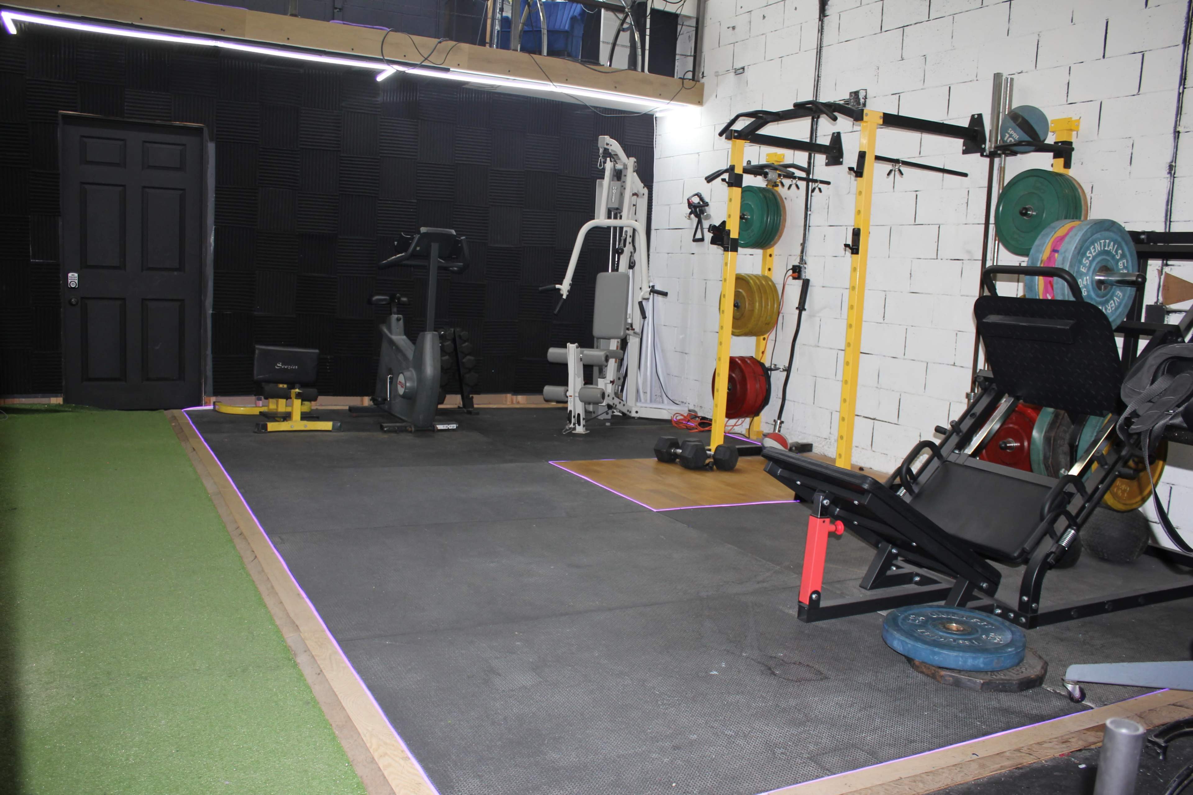An indoor gym space equipped with weightlifting and cardio machines, a padded floor, and various exercise weights.