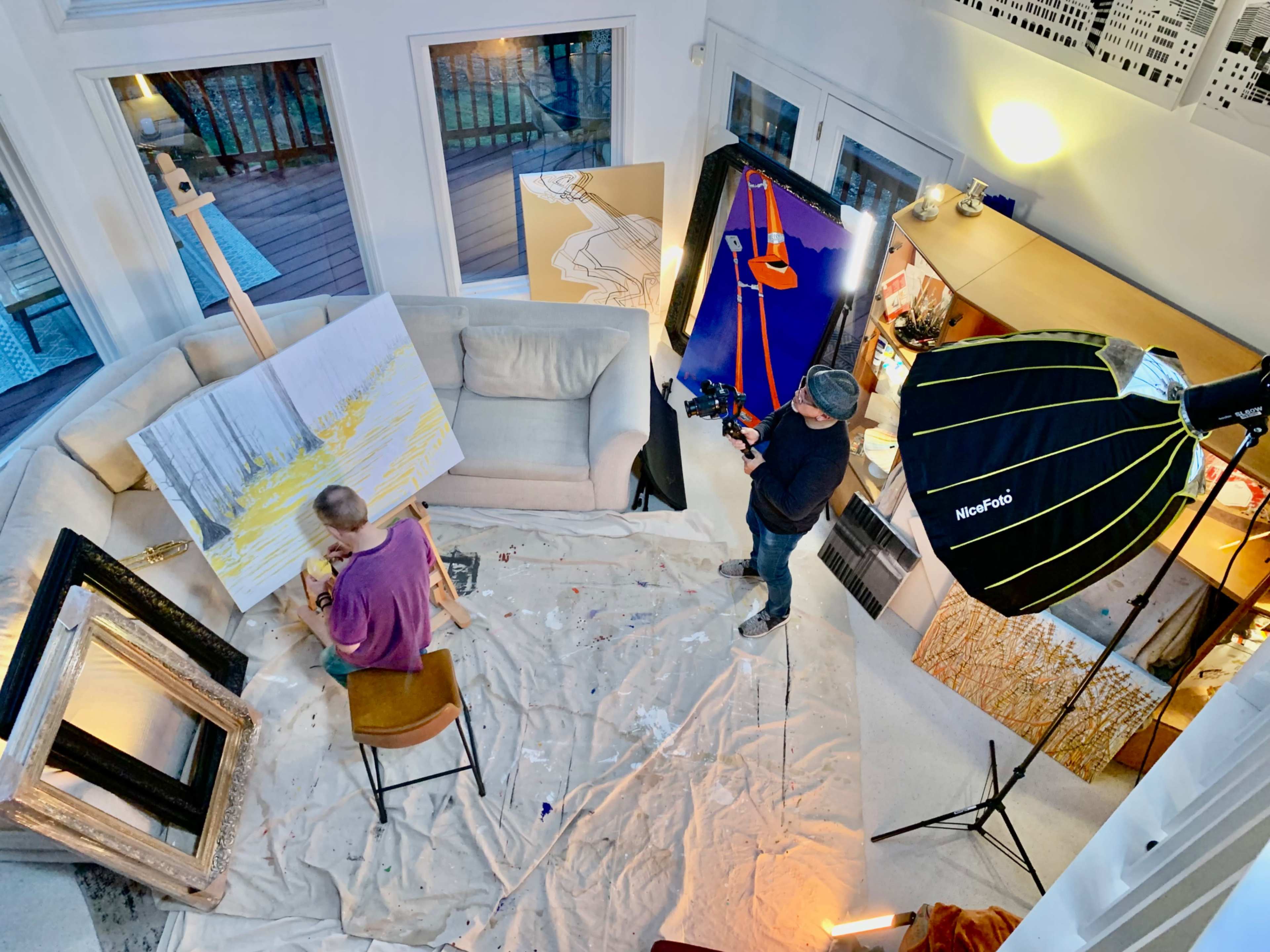 An artist is painting on a large canvas while a person records the process with a camera in a well-lit studio surrounded by various artworks and props.