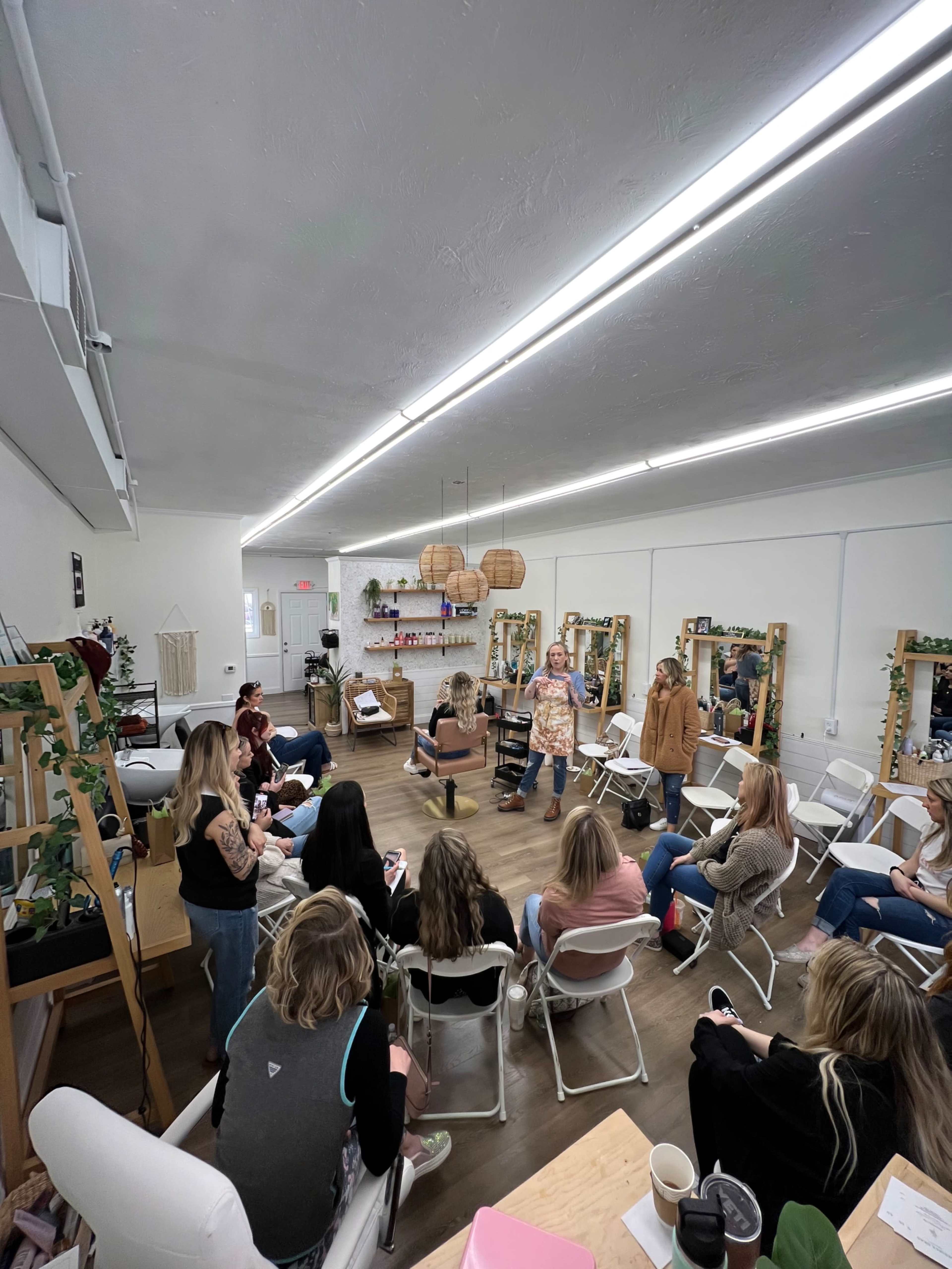 A group of people is seated in a well-lit room with mirrors and plants, while one person stands and speaks to them.