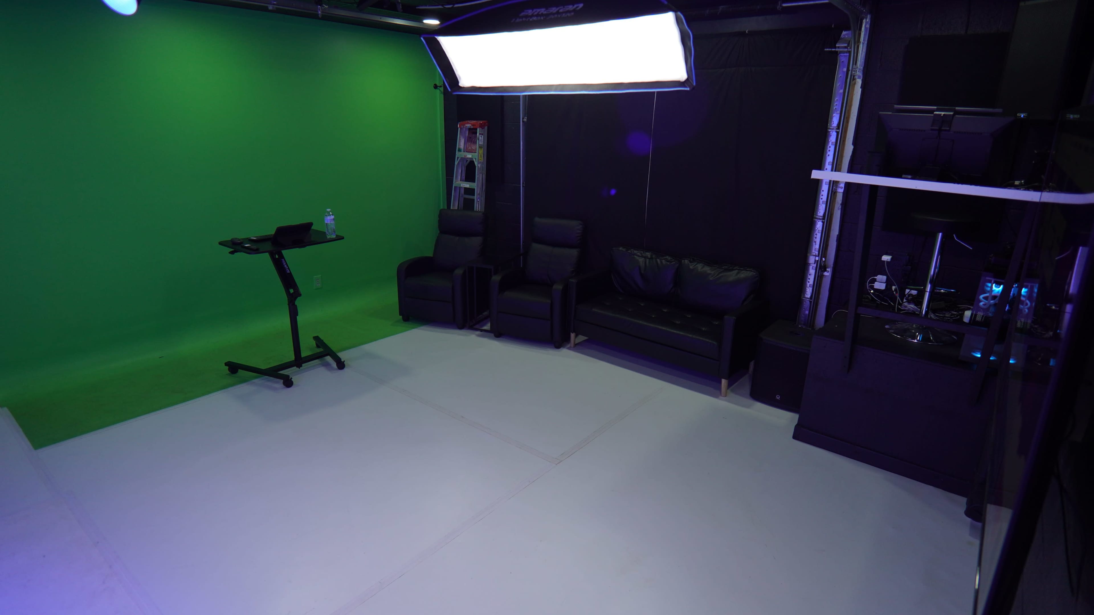The image shows a studio space with a green screen on one wall, black furniture including two chairs and a couch, and lighting equipment overhead.