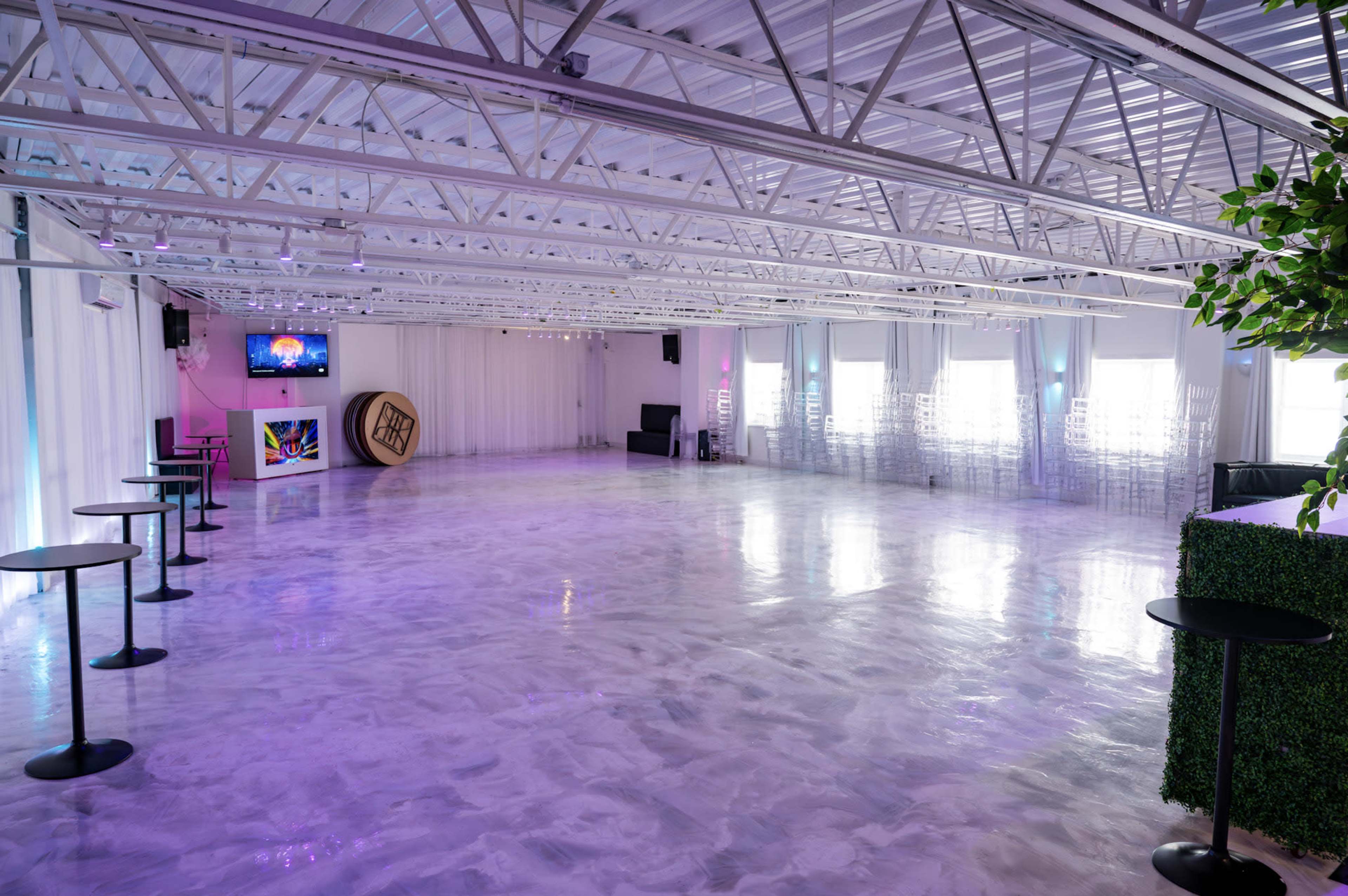 The image shows a spacious, modern event venue with polished floors, white walls, and a few sparse black bar tables, illuminated by colorful lights.