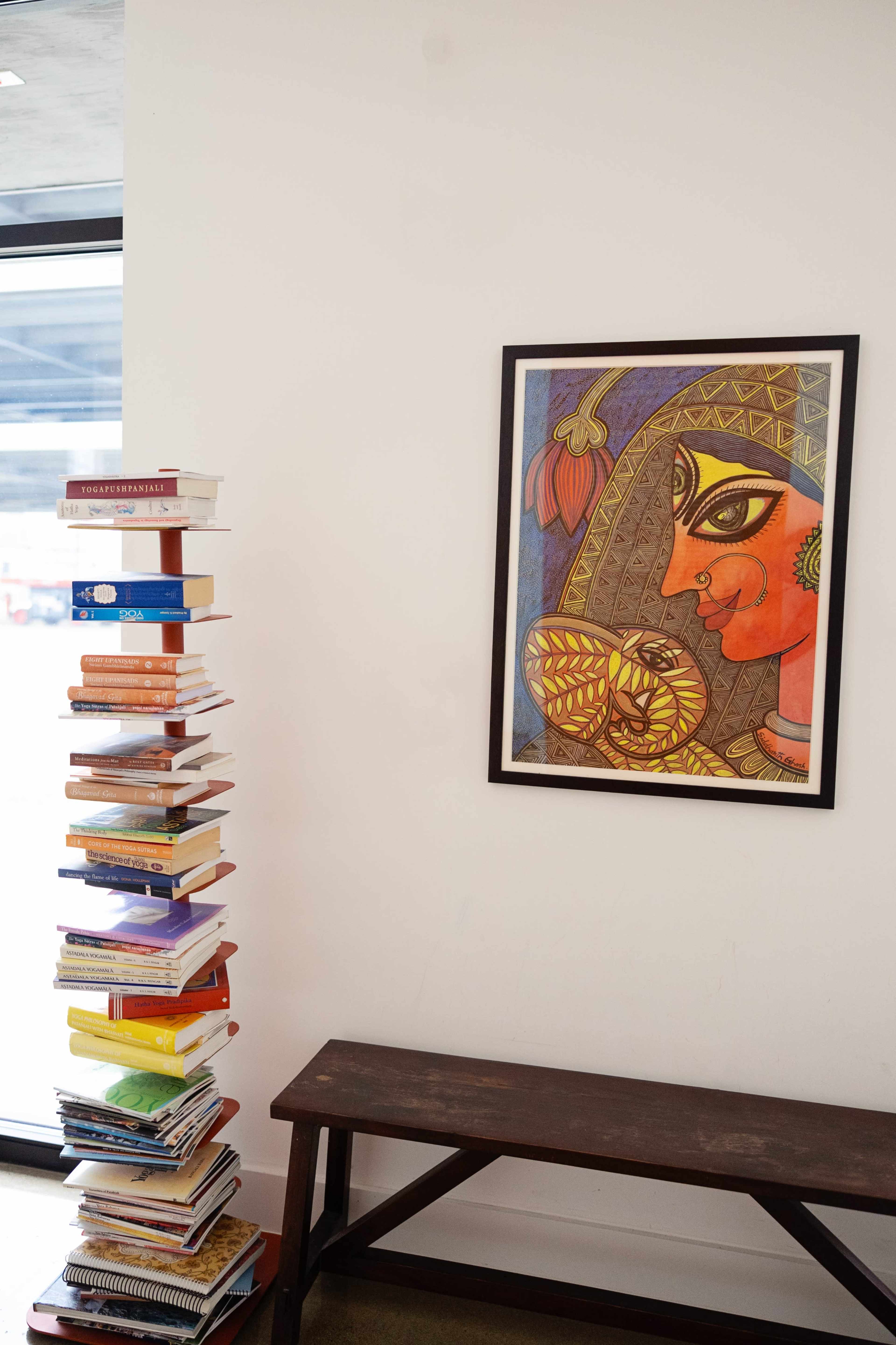 A stack of colorful books stands next to a framed painting of a woman with traditional Indian attire on a wall.