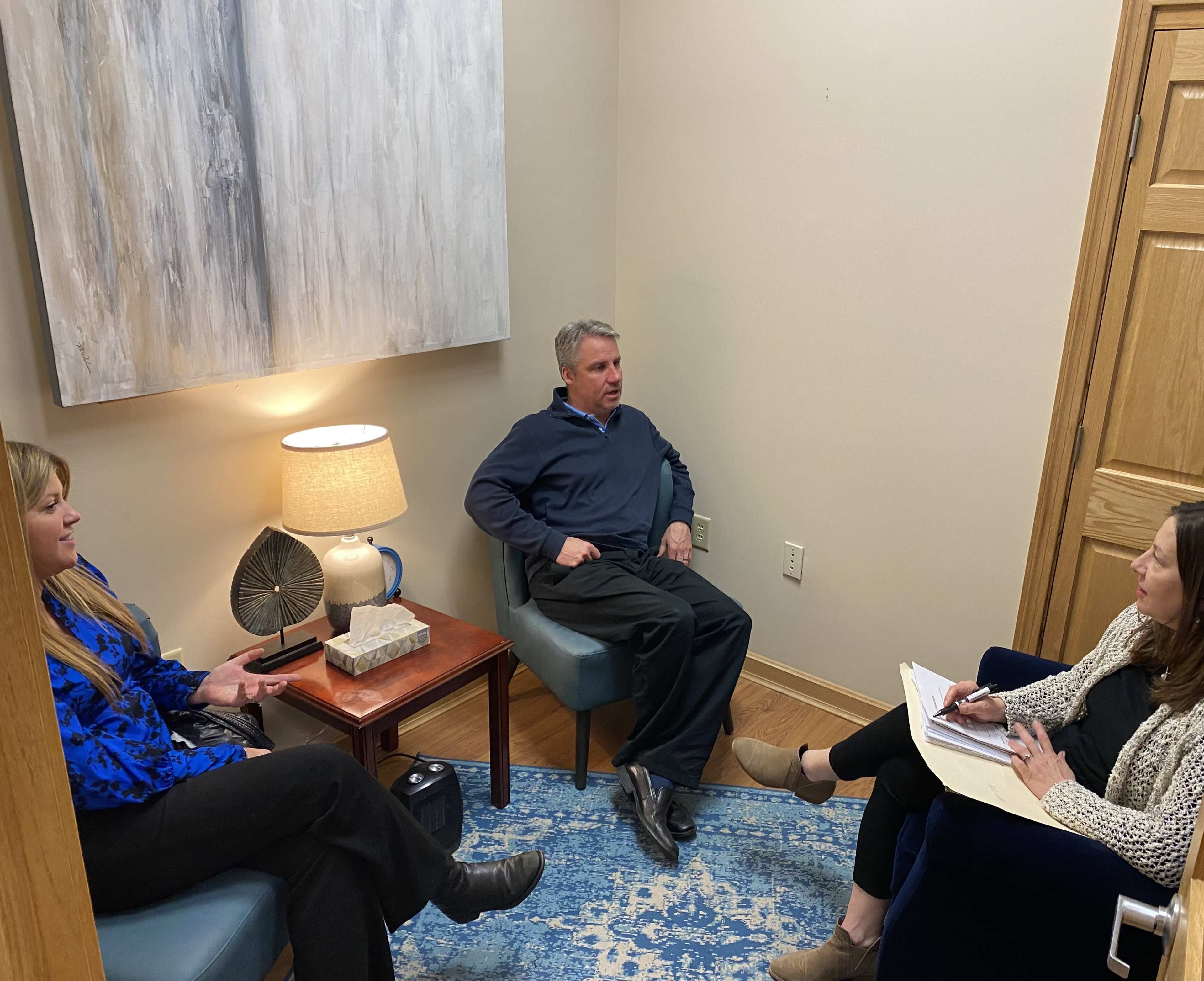 Three people sit in a small, well-lit room, engaging in a discussion, with a painting and a lamp on a side table nearby.