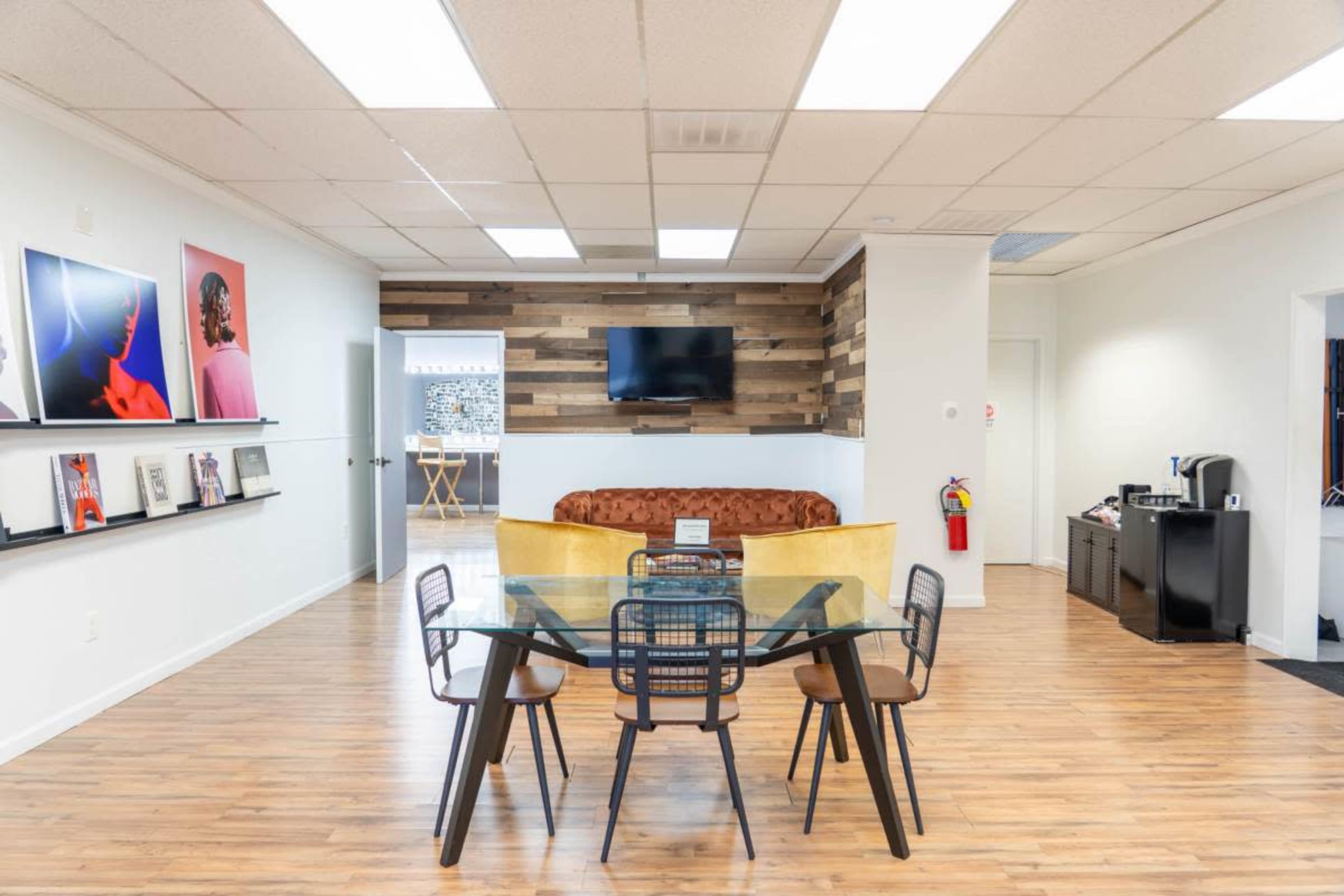 The image shows a modern office interior featuring a glass dining table with black chairs, a brown sofa, and artwork on the walls, along with a television and wooden paneling.
