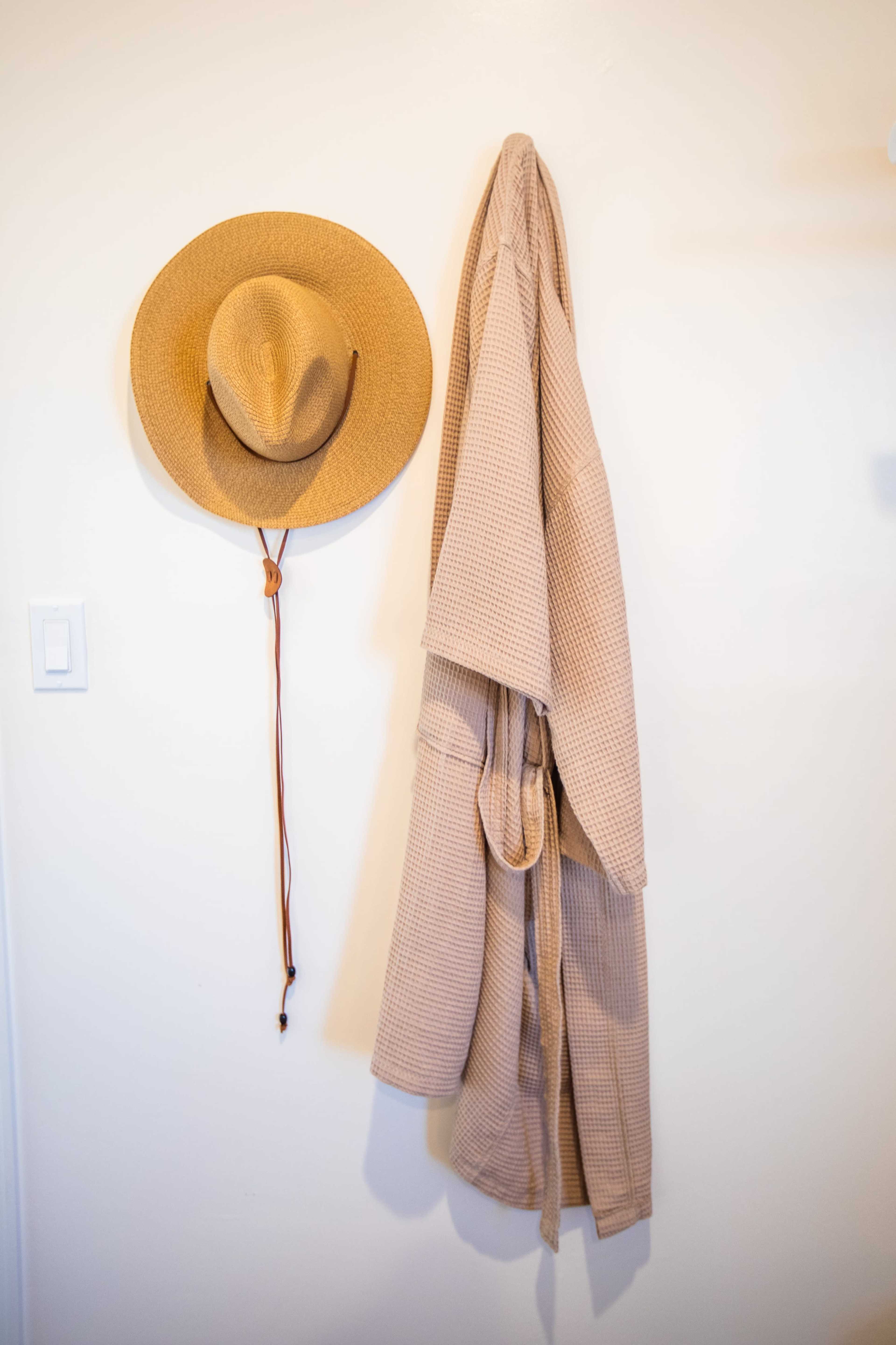 A woven straw hat and a beige bathrobe are hung on a white wall.
