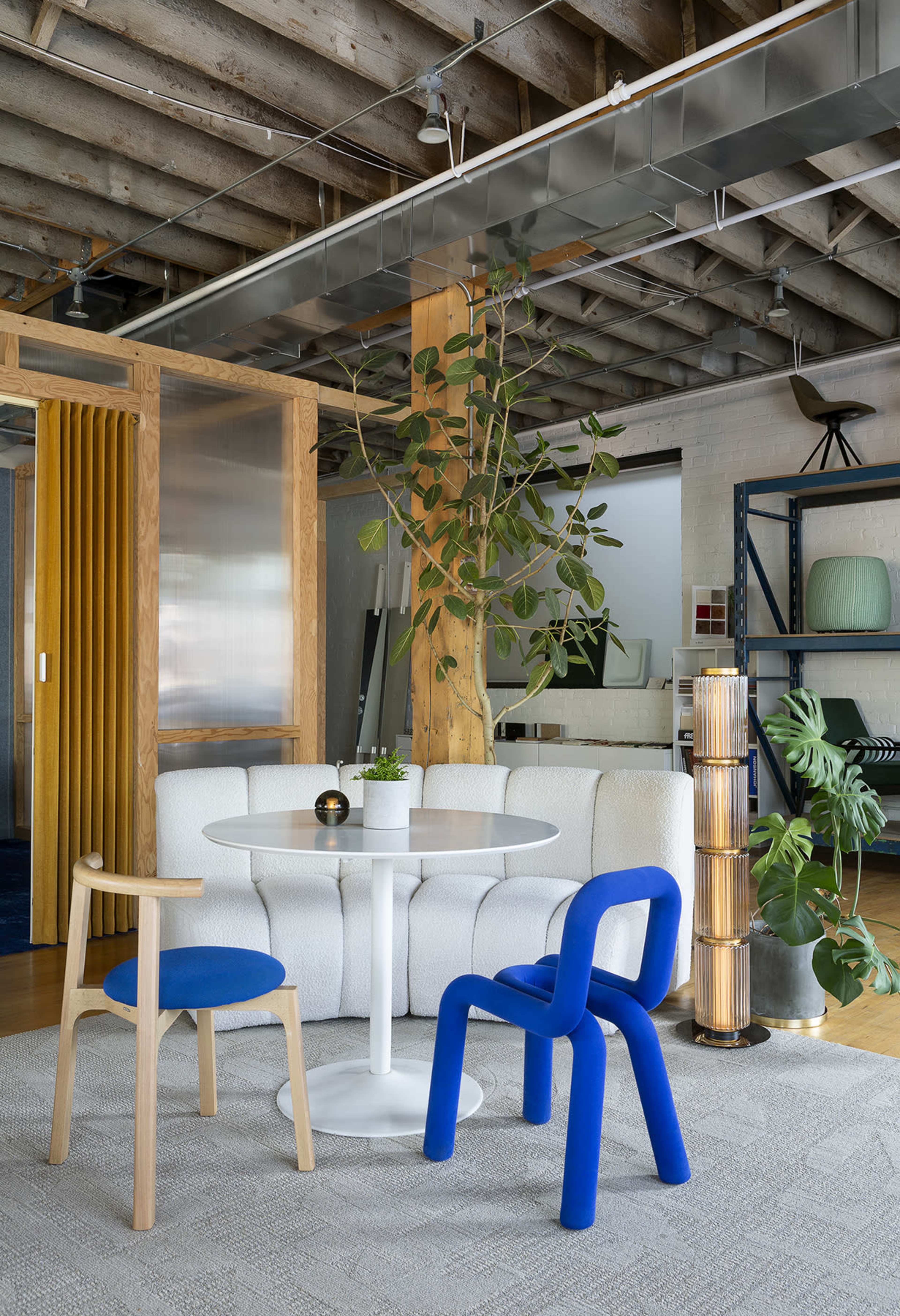The image shows a modern interior space featuring a circular white table surrounded by two colorful chairs, a potted plant, and a textured area rug, with wooden beams and metal accents in the background.