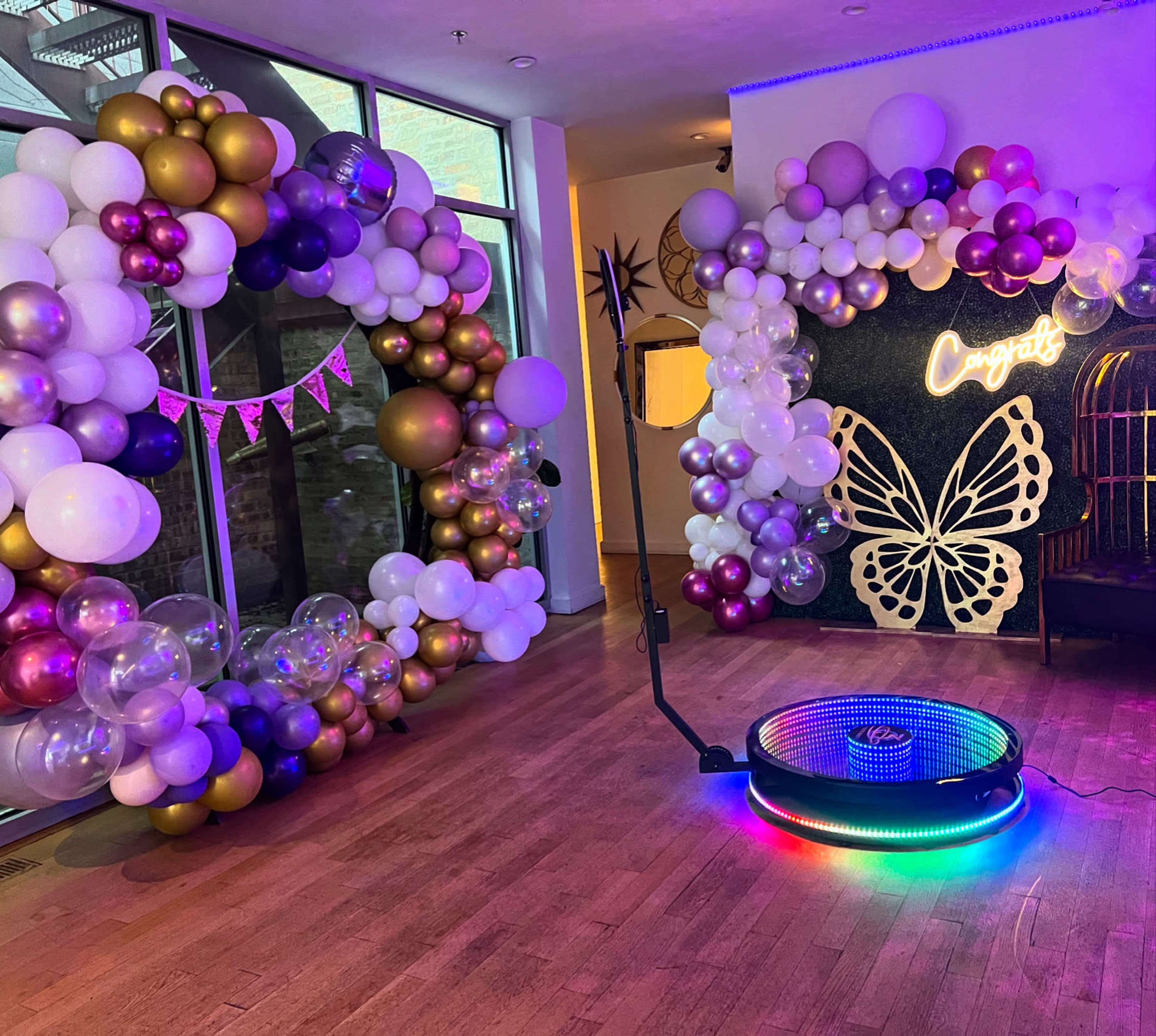 The scene features a decorated event space with two large balloon arches in purple, gold, and white, along with a light-up circular platform and a butterfly-themed backdrop.