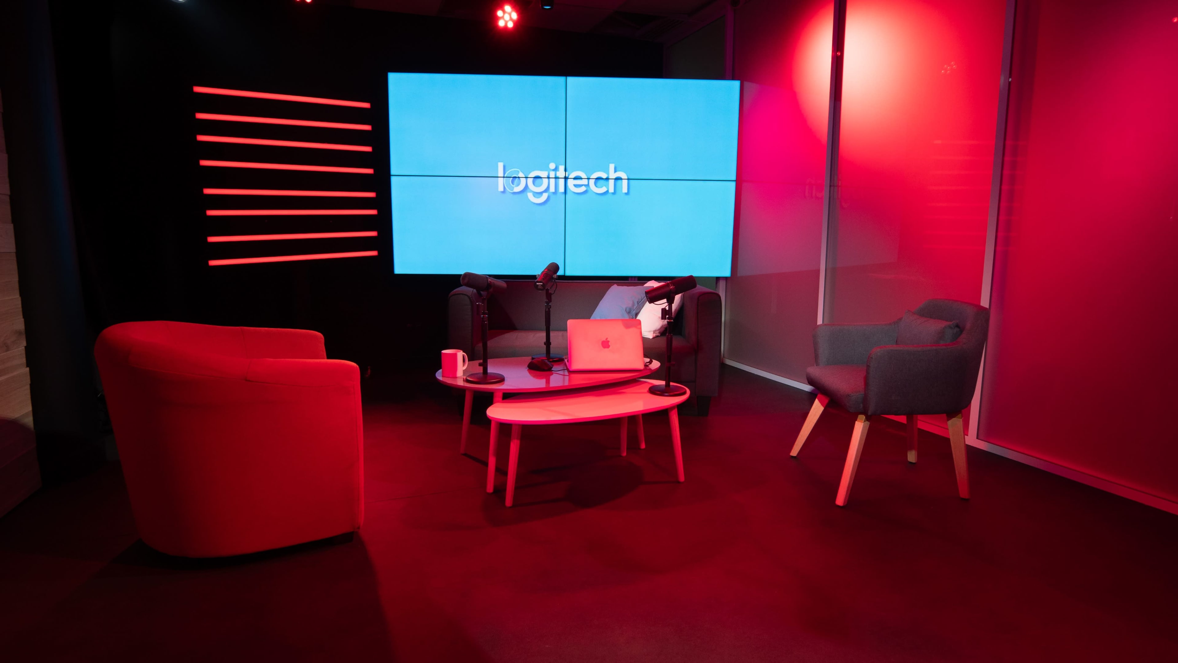 A modern studio setting features red-accented furniture, a large screen displaying the Logitech logo, and equipment for a podcast or presentation.