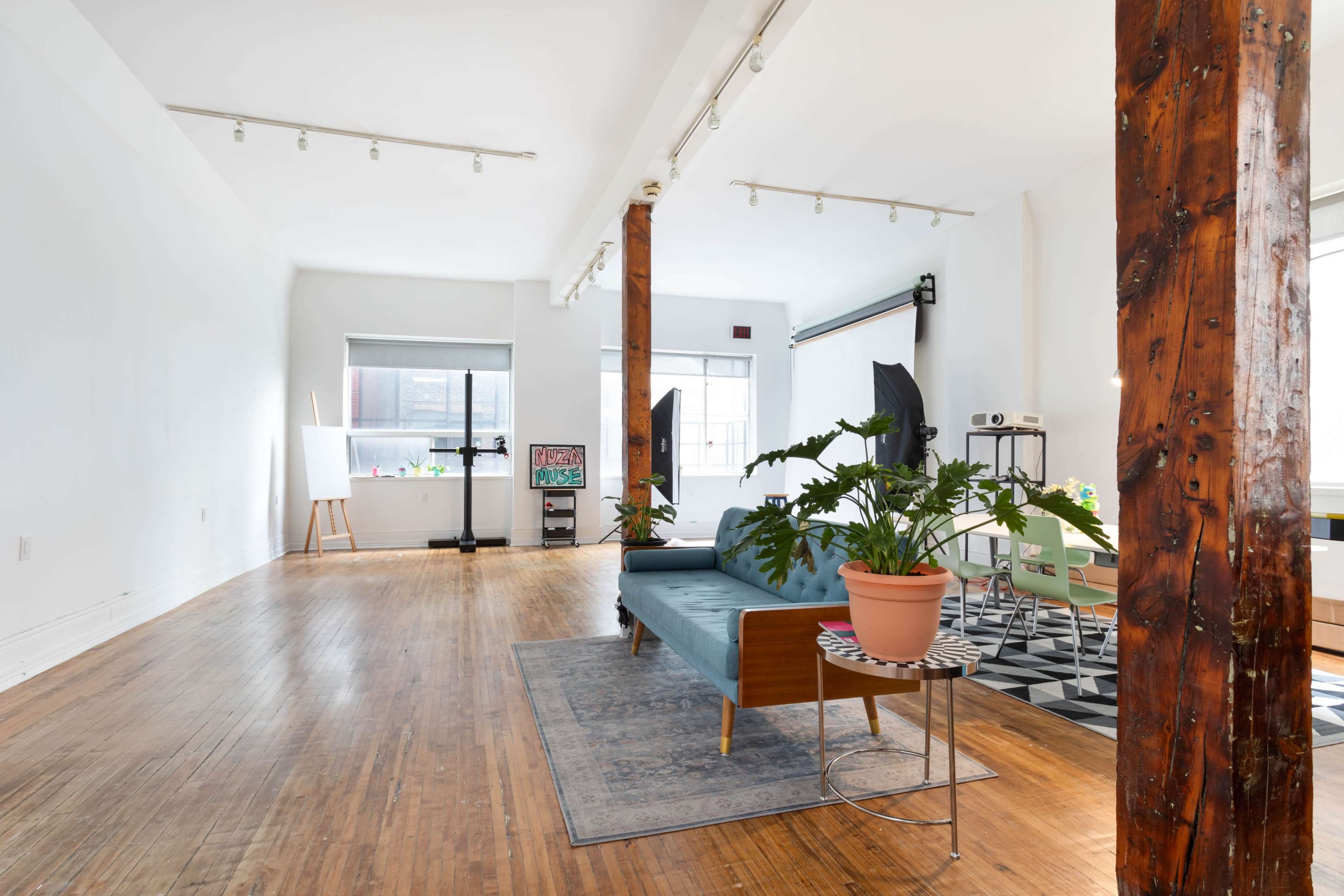 A spacious, well-lit room features wooden pillars, a blue sofa, a potted plant on a small table, and various furniture arrangements.