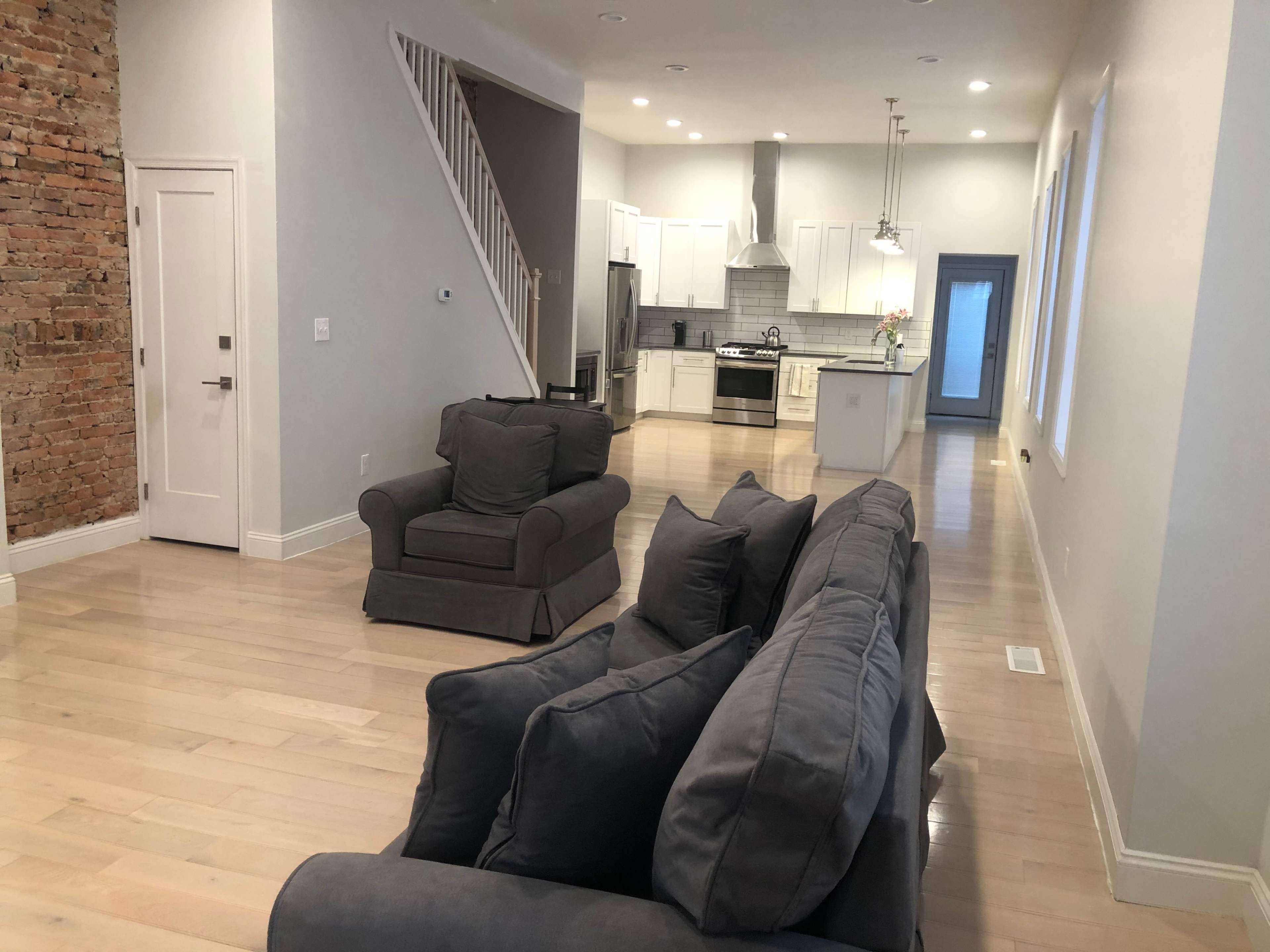 A modern living room and kitchen area with hardwood floors, exposed brick walls, and a gray sofa facing an open kitchen.