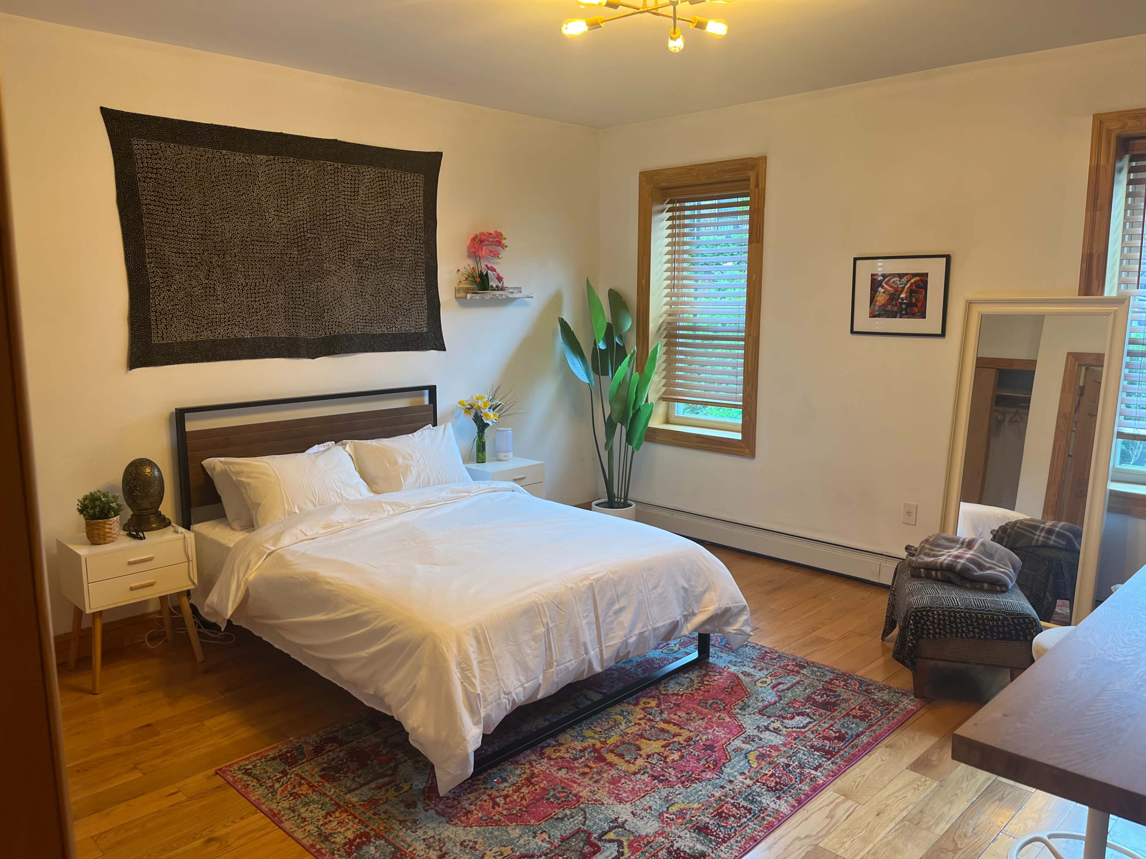 A tidy bedroom featuring a bed with white bedding, a patterned area rug, a wooden dresser, a mirror, and large windows with wooden blinds.