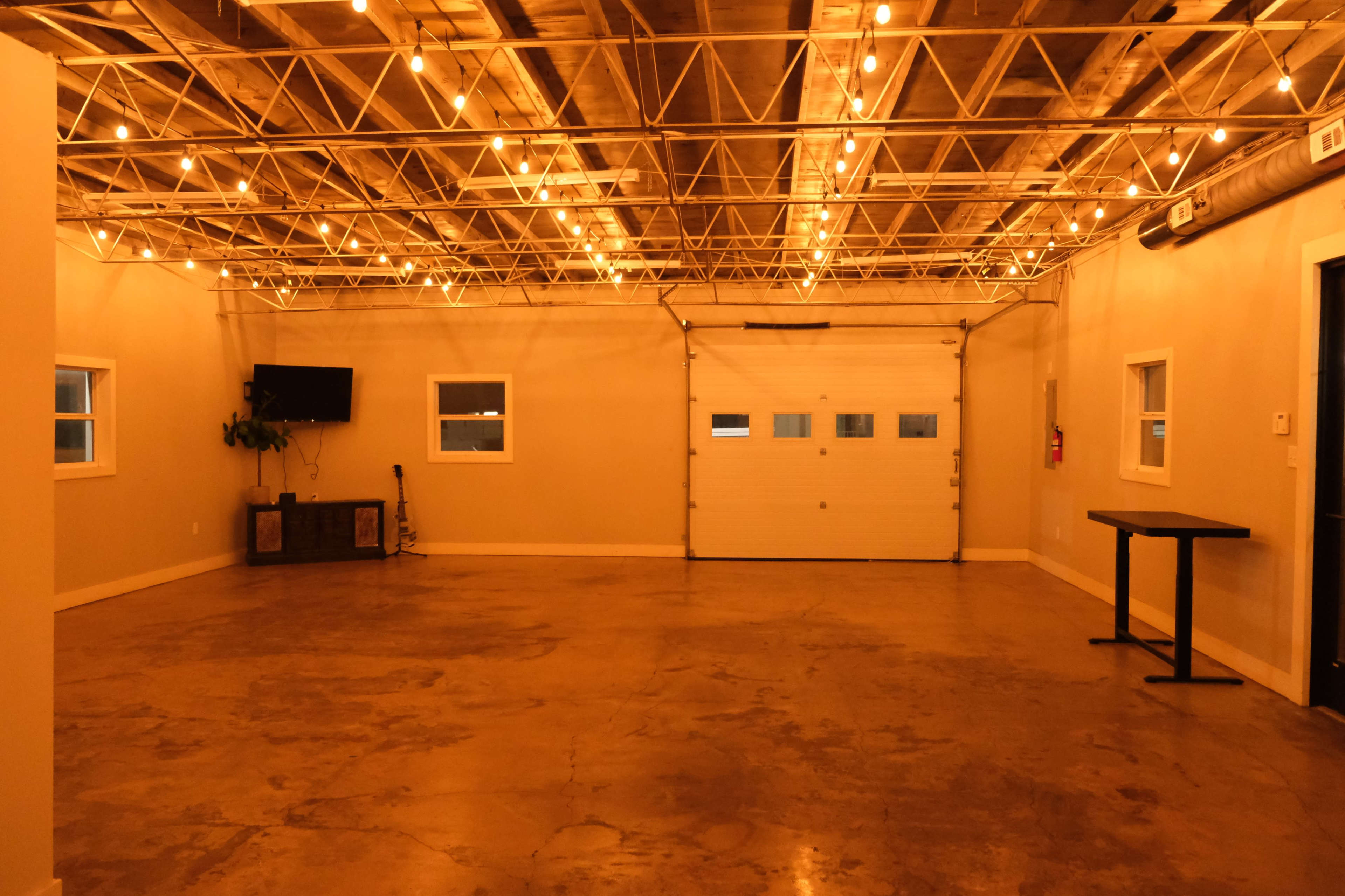 A spacious, well-lit garage with a polished concrete floor, string lights overhead, a wall-mounted television, and an overhead door on one side.