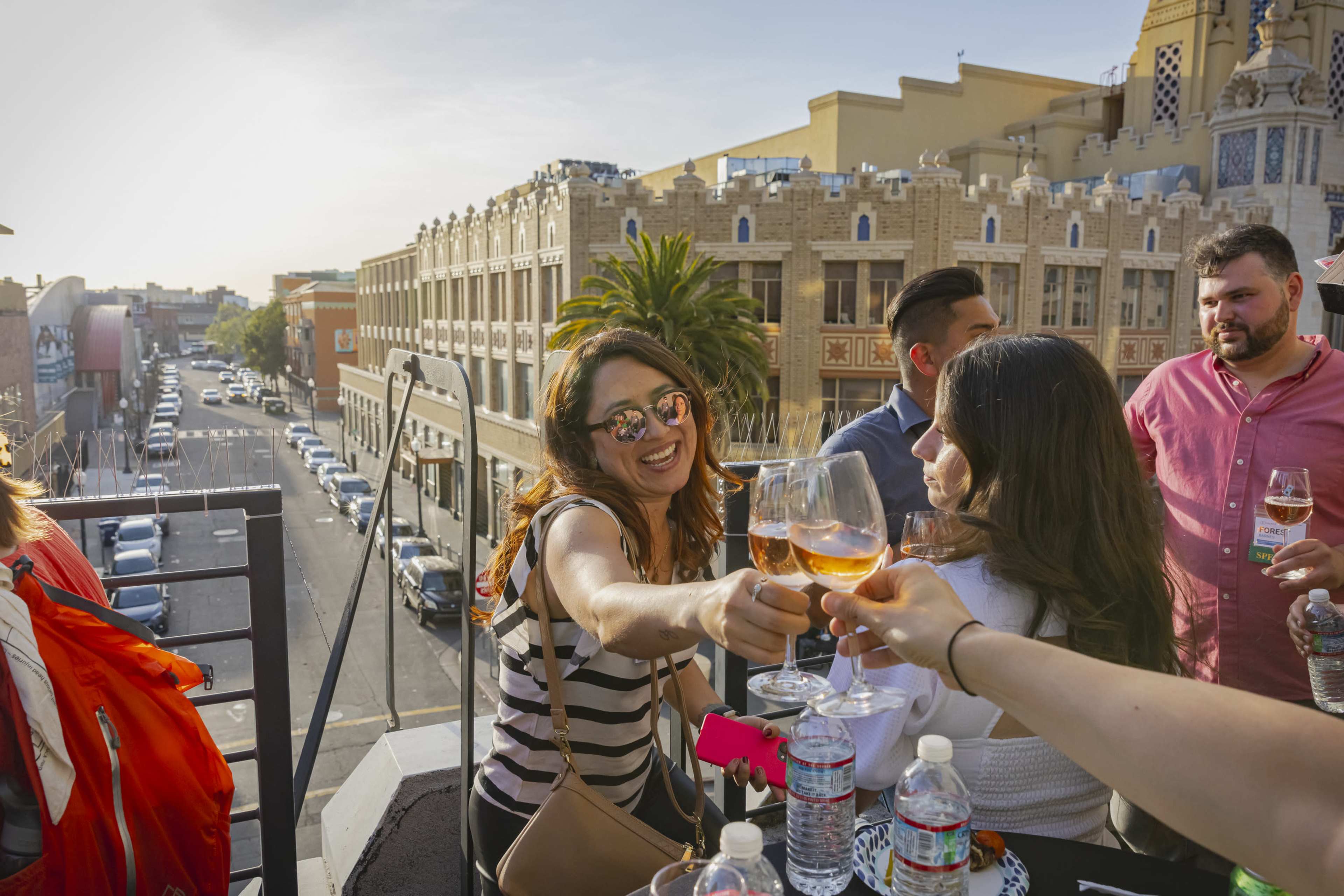 A group of people toasts with glasses of wine on a rooftop terrace overlooking a city street at sunset.