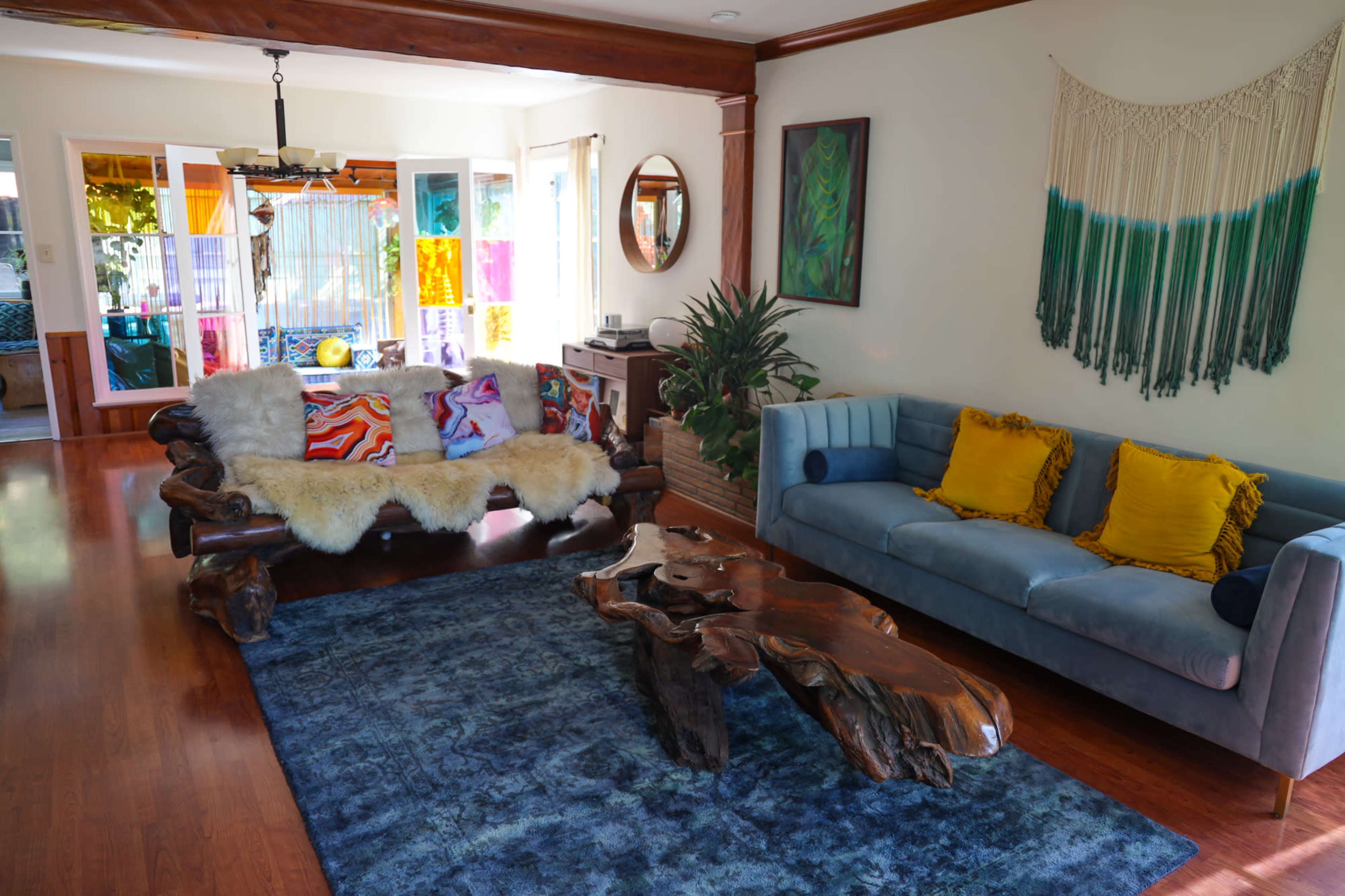 The living room features a wooden coffee table, a blue couch, a colorful patterned sofa adorned with cushions, and a large area rug on polished wood floors.
