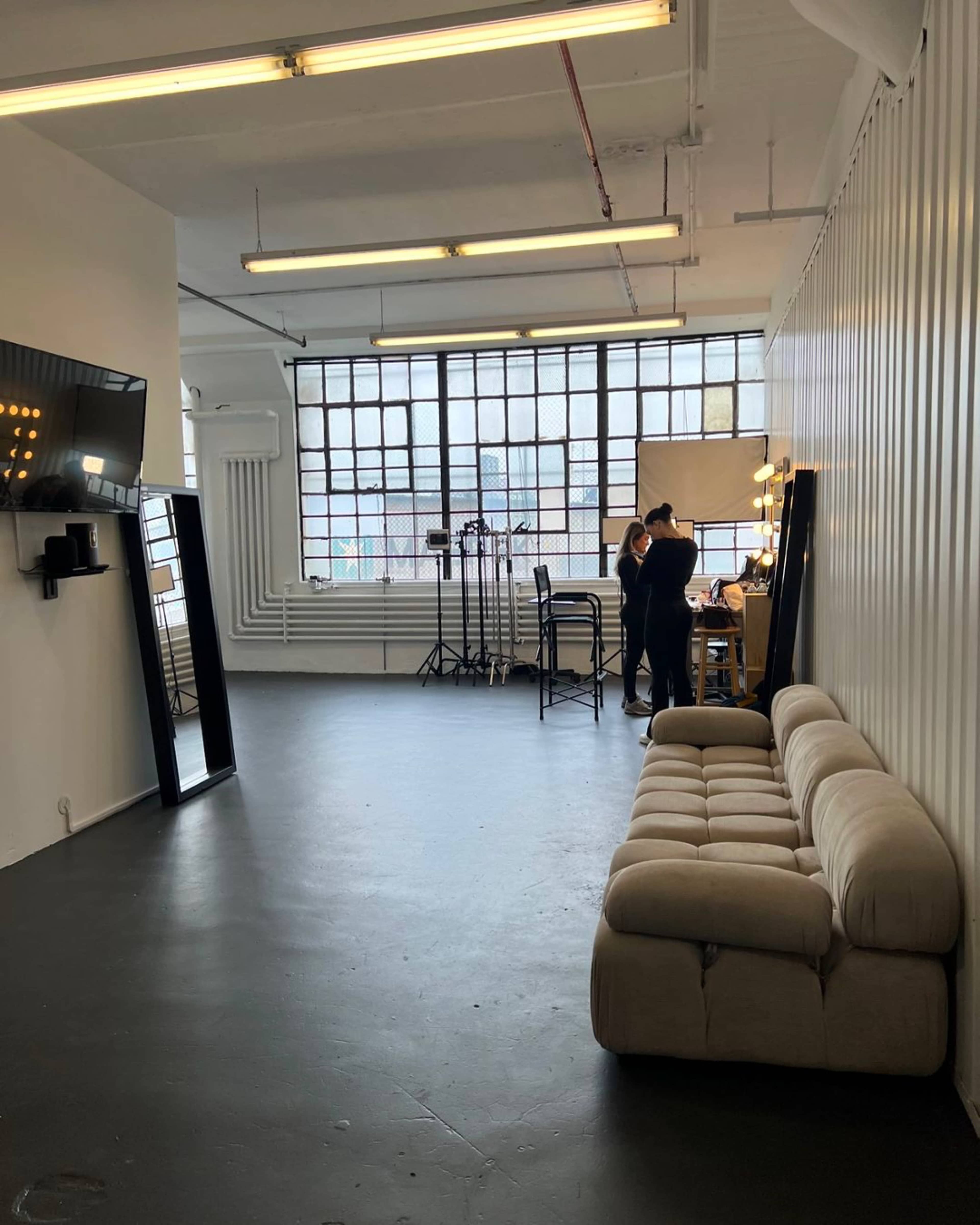 A spacious, minimalistic studio with a beige sofa, large windows, and two individuals working at a table with lighting equipment in the background.