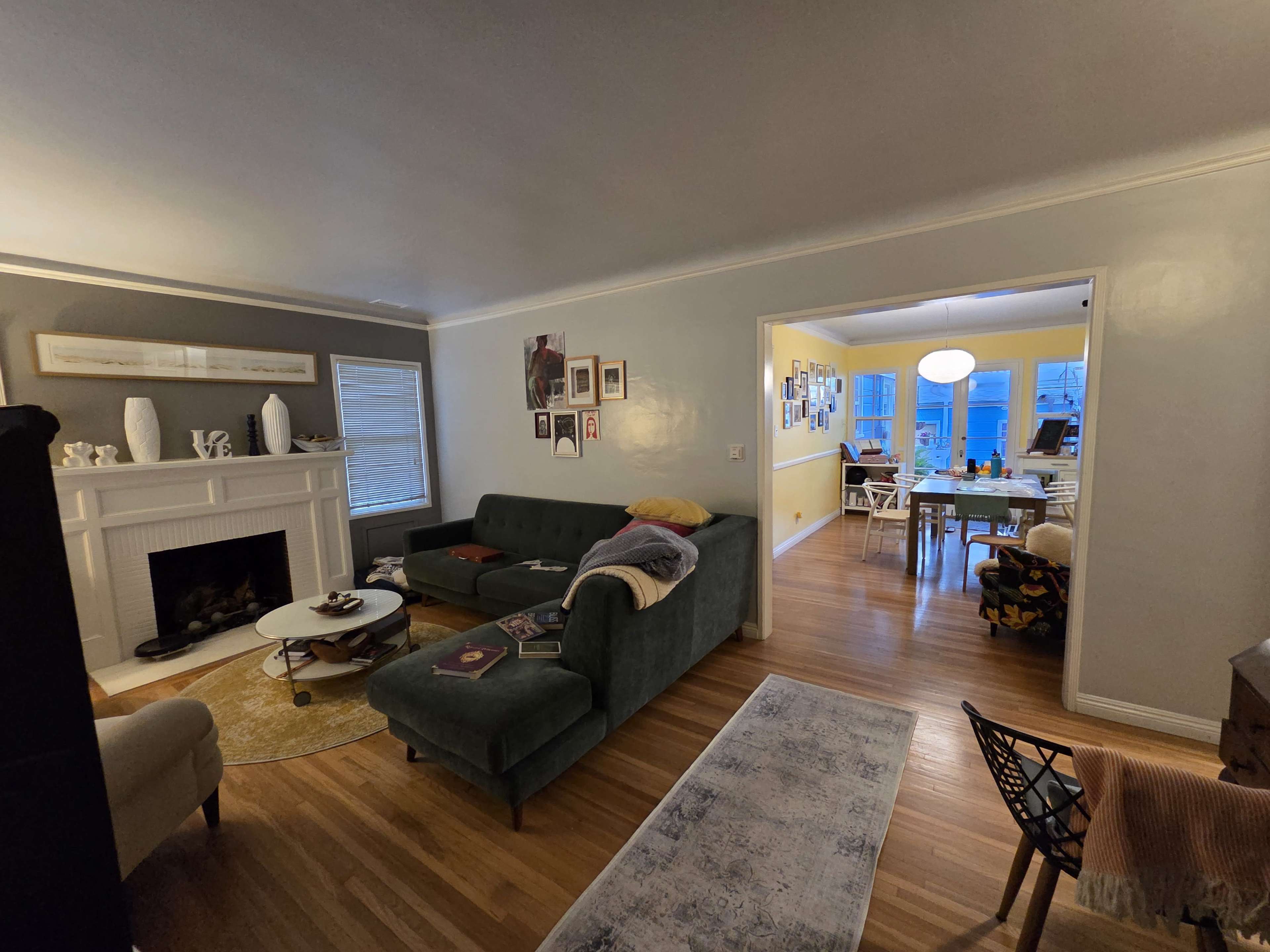 The image shows a living room with a green sectional sofa, a coffee table, and a fireplace, leading into a dining area with a table illuminated by overhead lighting.