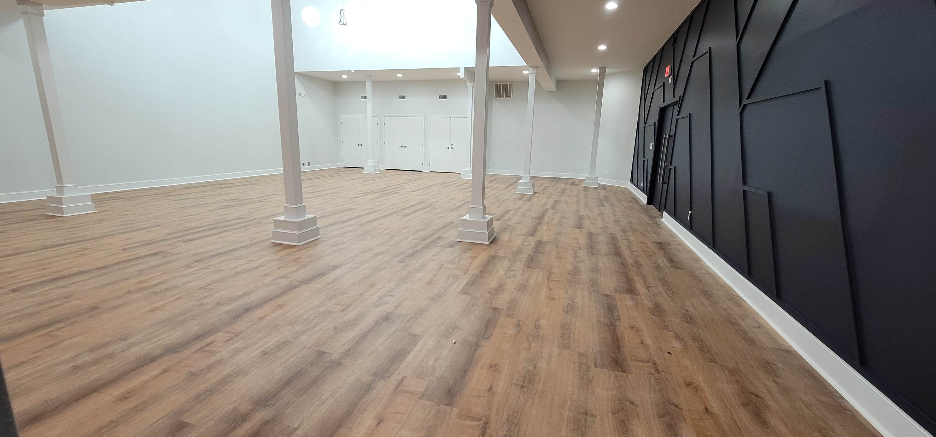 An expansive interior space features wooden flooring, white columns, and a dark accent wall with geometric designs.