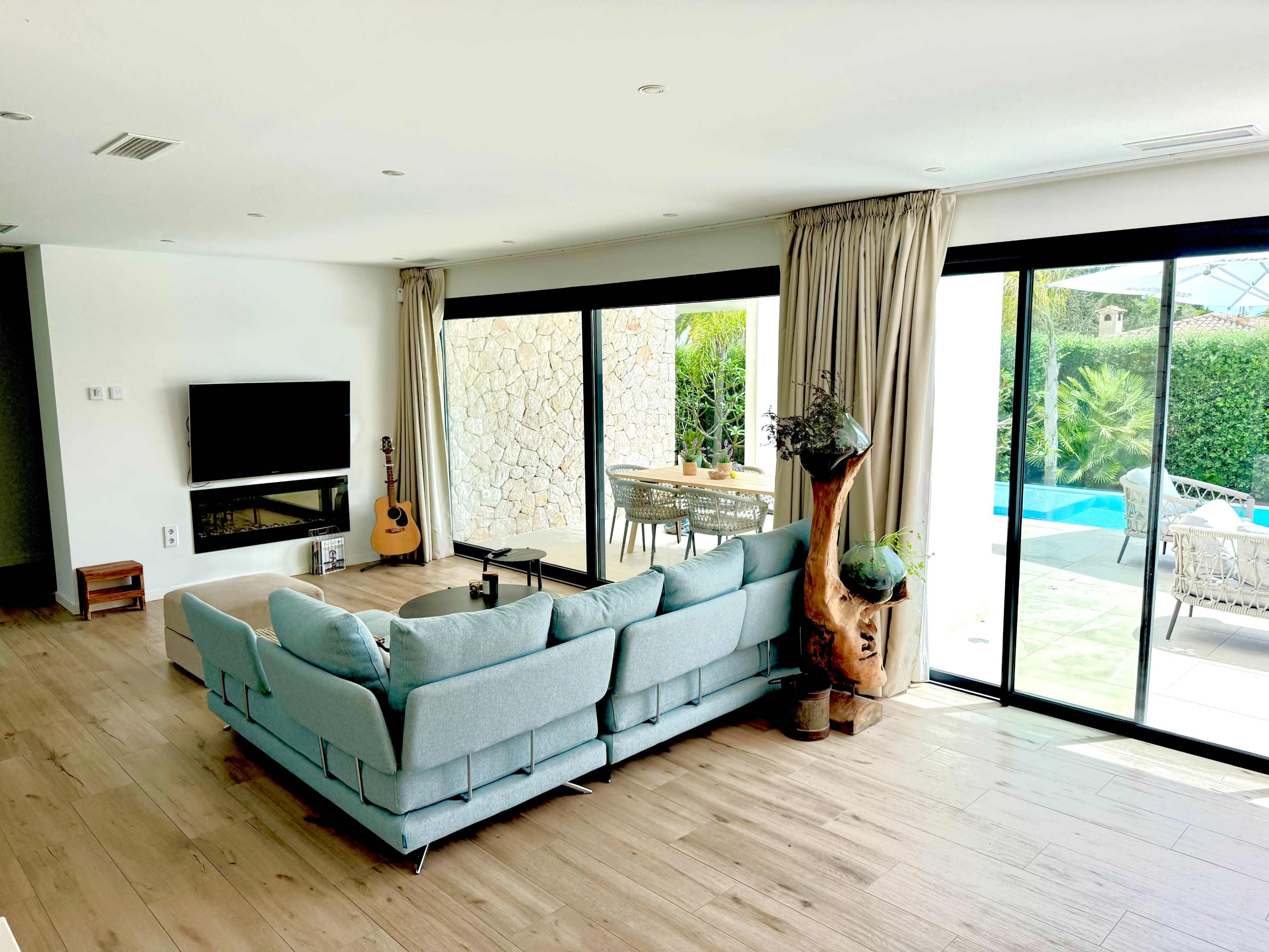 A modern living room features a light blue sectional sofa, a television mounted on the wall, and large glass doors that open to a pool area with outdoor seating.
