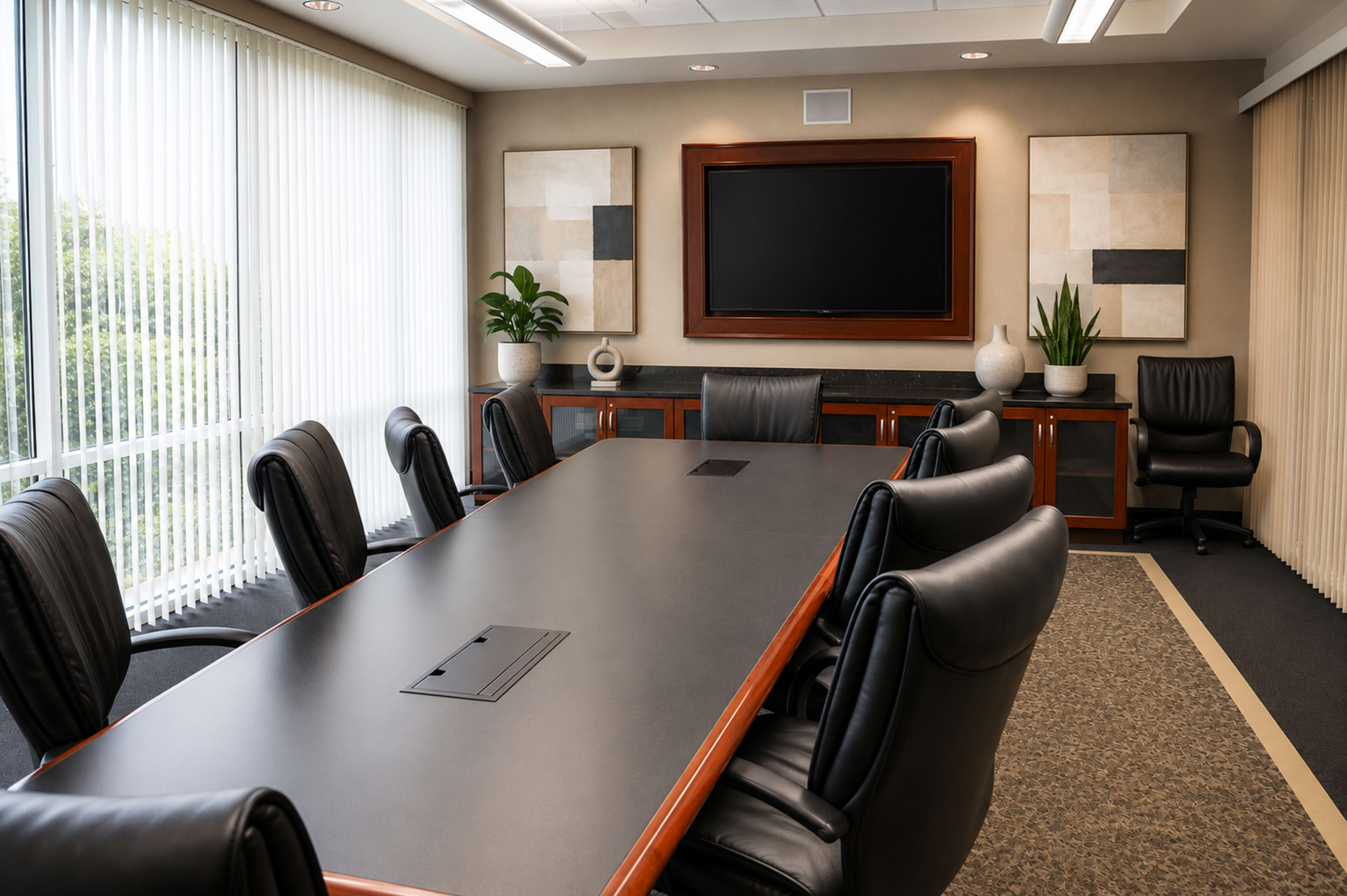 A modern conference room features a long black table surrounded by leather chairs, with large windows letting in natural light and abstract art on the walls.