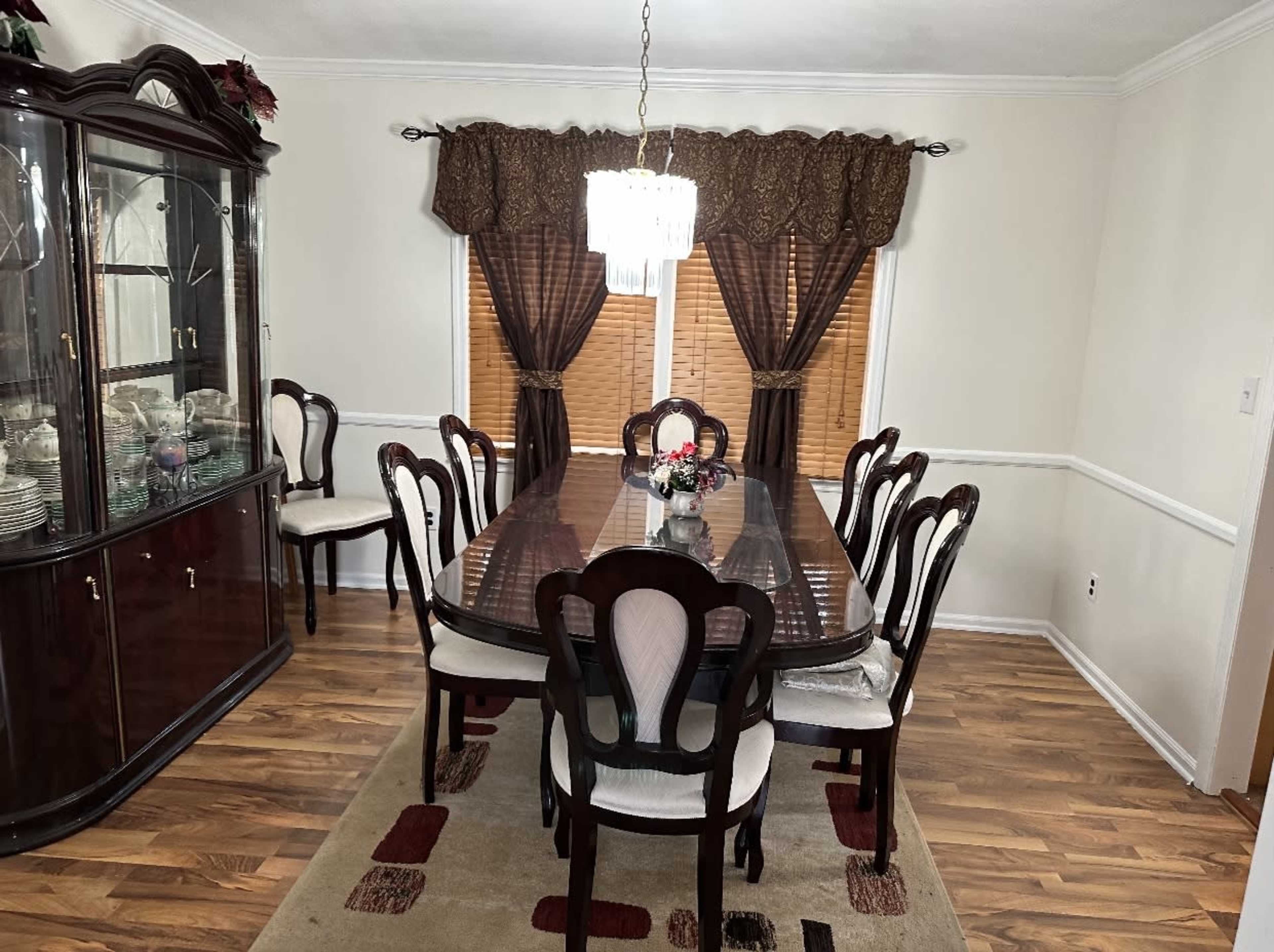 A dining room features a large table with eight chairs, a china cabinet, and windows dressed with dark curtains.