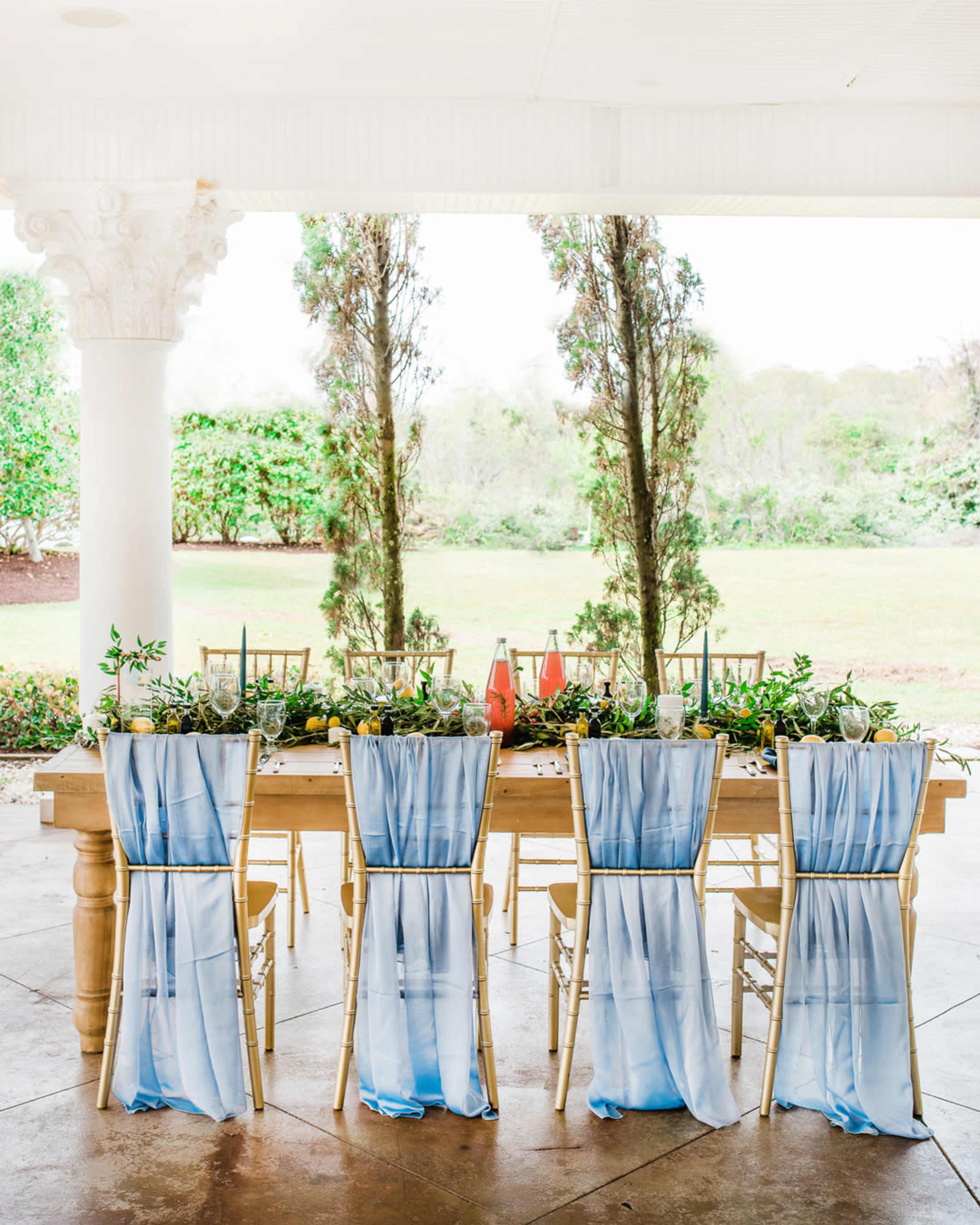 A rustic wooden table set for a meal features light blue fabric draped over the backs of four chairs, with greenery and decorative items arranged along the center, all under a covered porch.