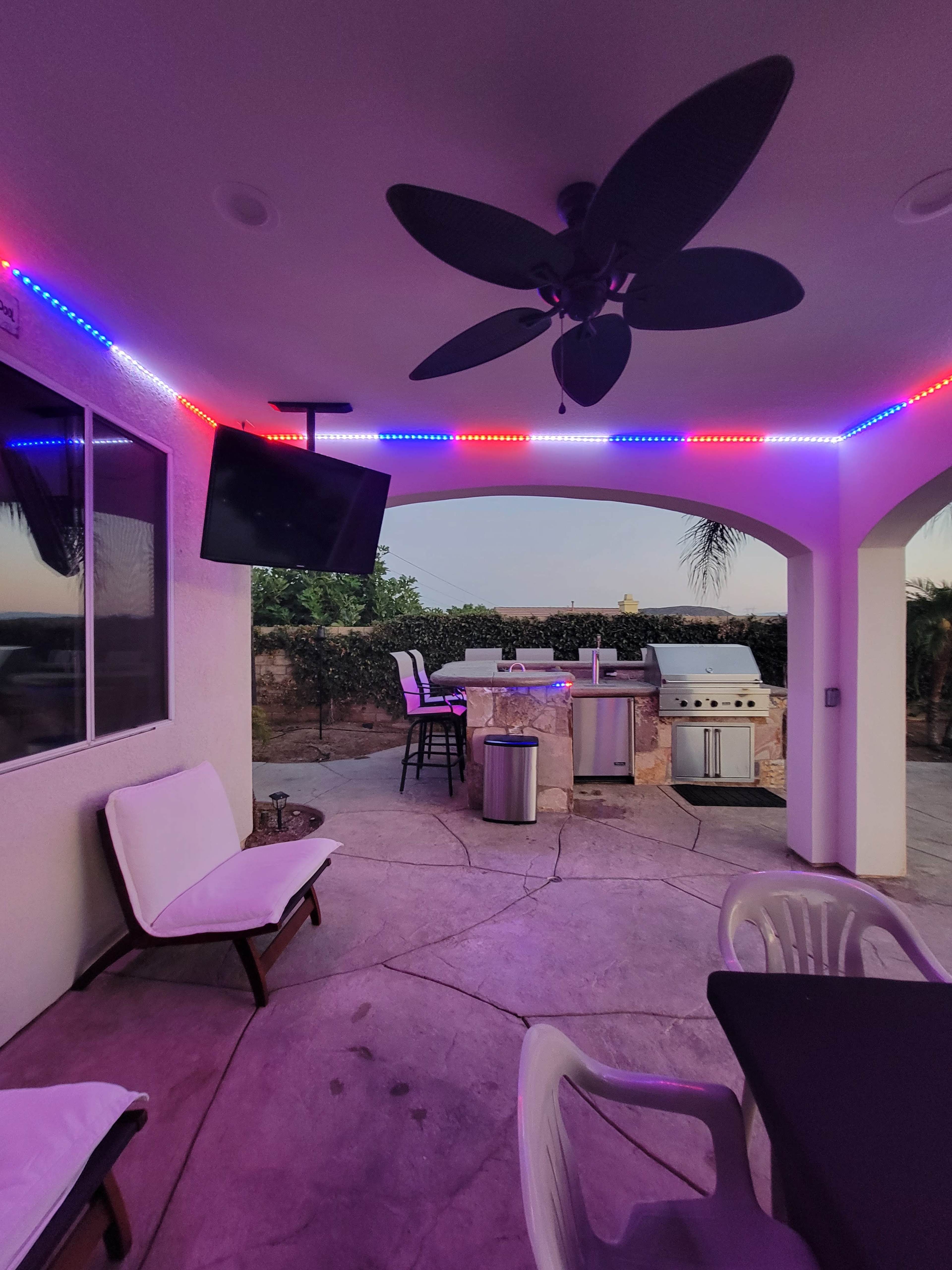 A covered outdoor patio area with a grill, refrigerator, and seating, illuminated by multicolored LED lights.