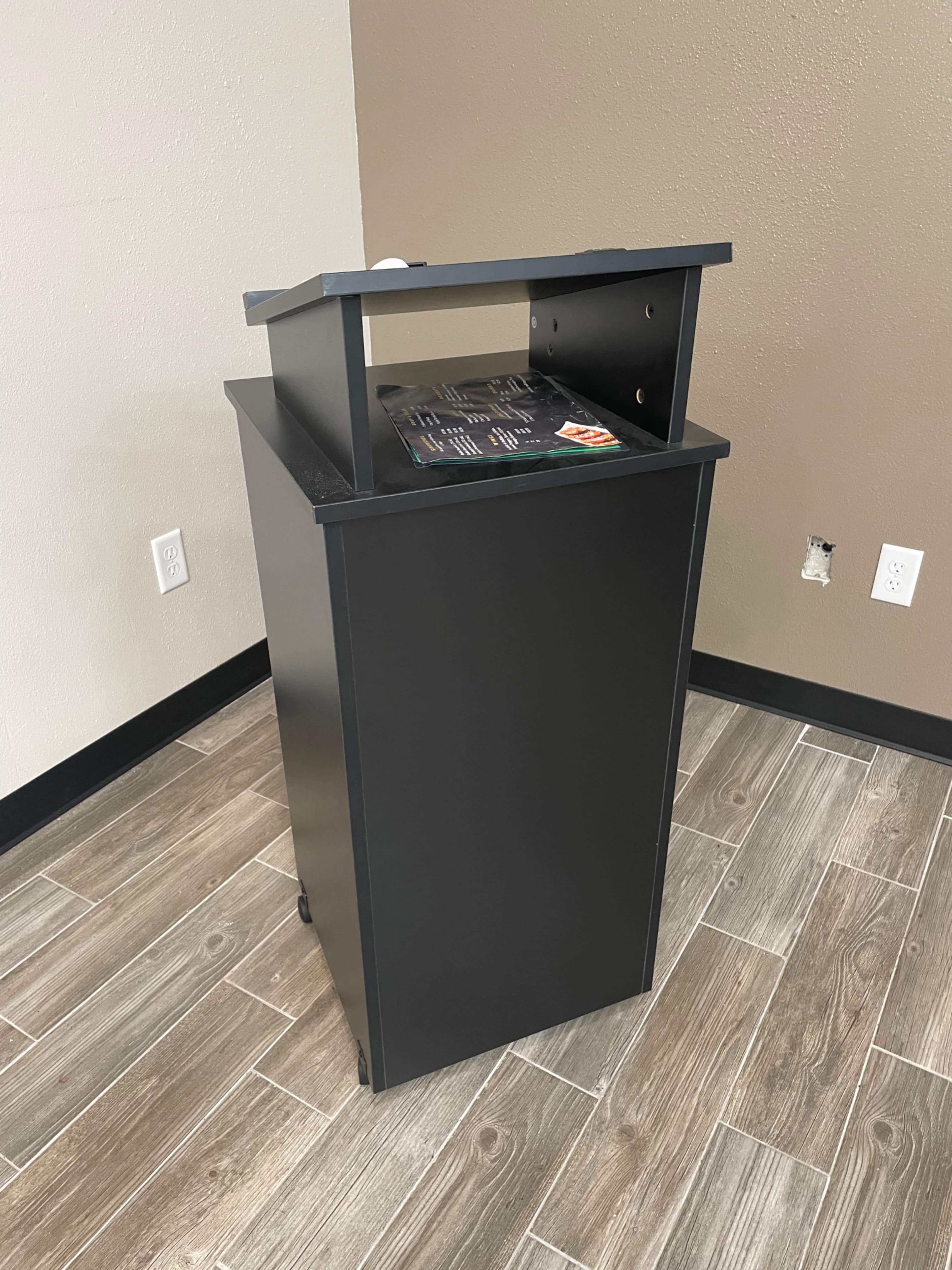 A black podium with a flat top and wheels is positioned on a wooden floor against a plain wall.