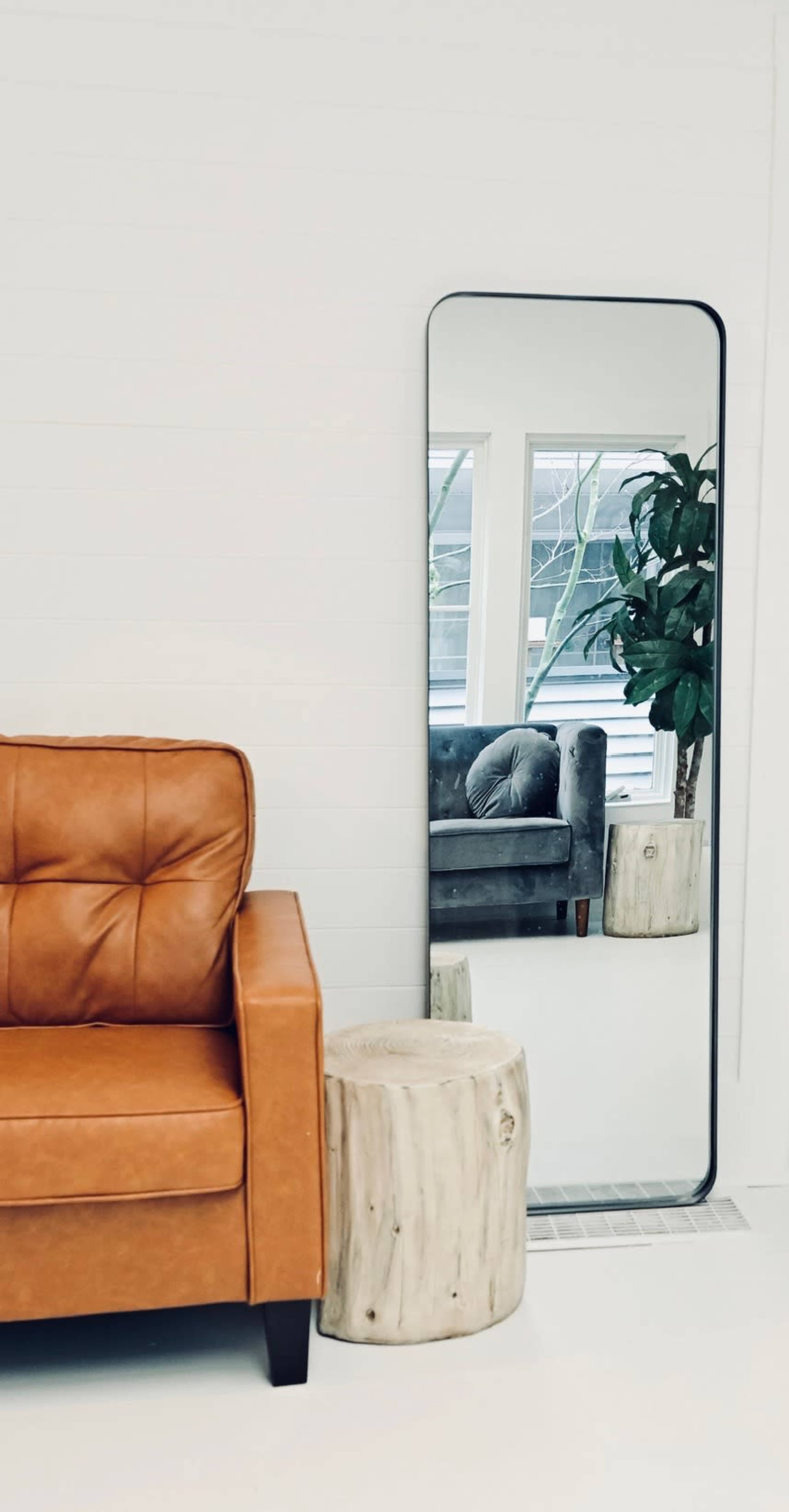 The image features a brown leather couch next to a round wooden side table, with a tall mirror reflecting a plant and another couch in the background.