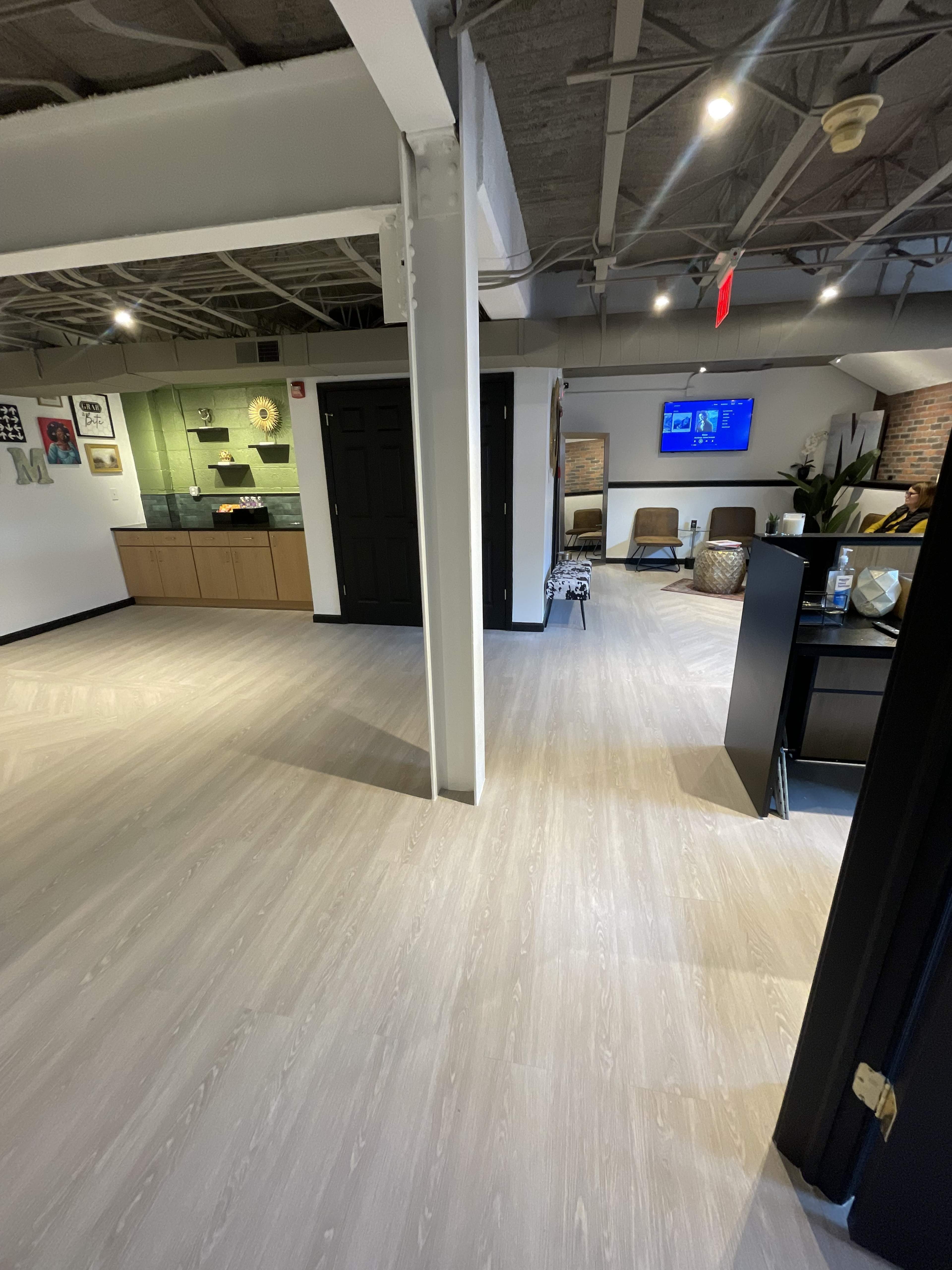 The image shows an interior space featuring a modern design with light-colored flooring, a kitchenette area, and seating arranged in a lounge-like setup.