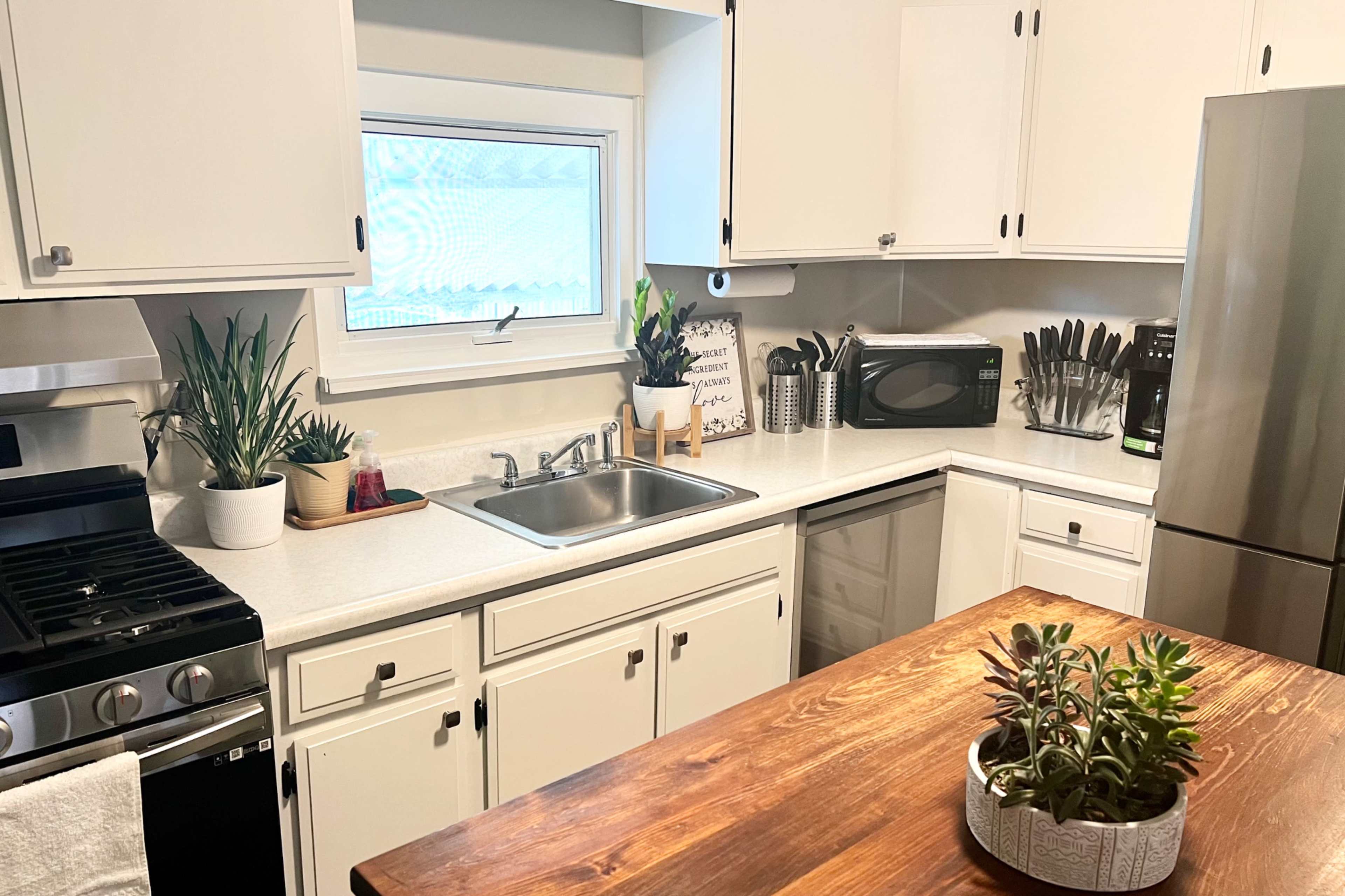 A bright kitchen features white cabinets, a stainless steel refrigerator, a sink, and a wooden island with potted succulents.
