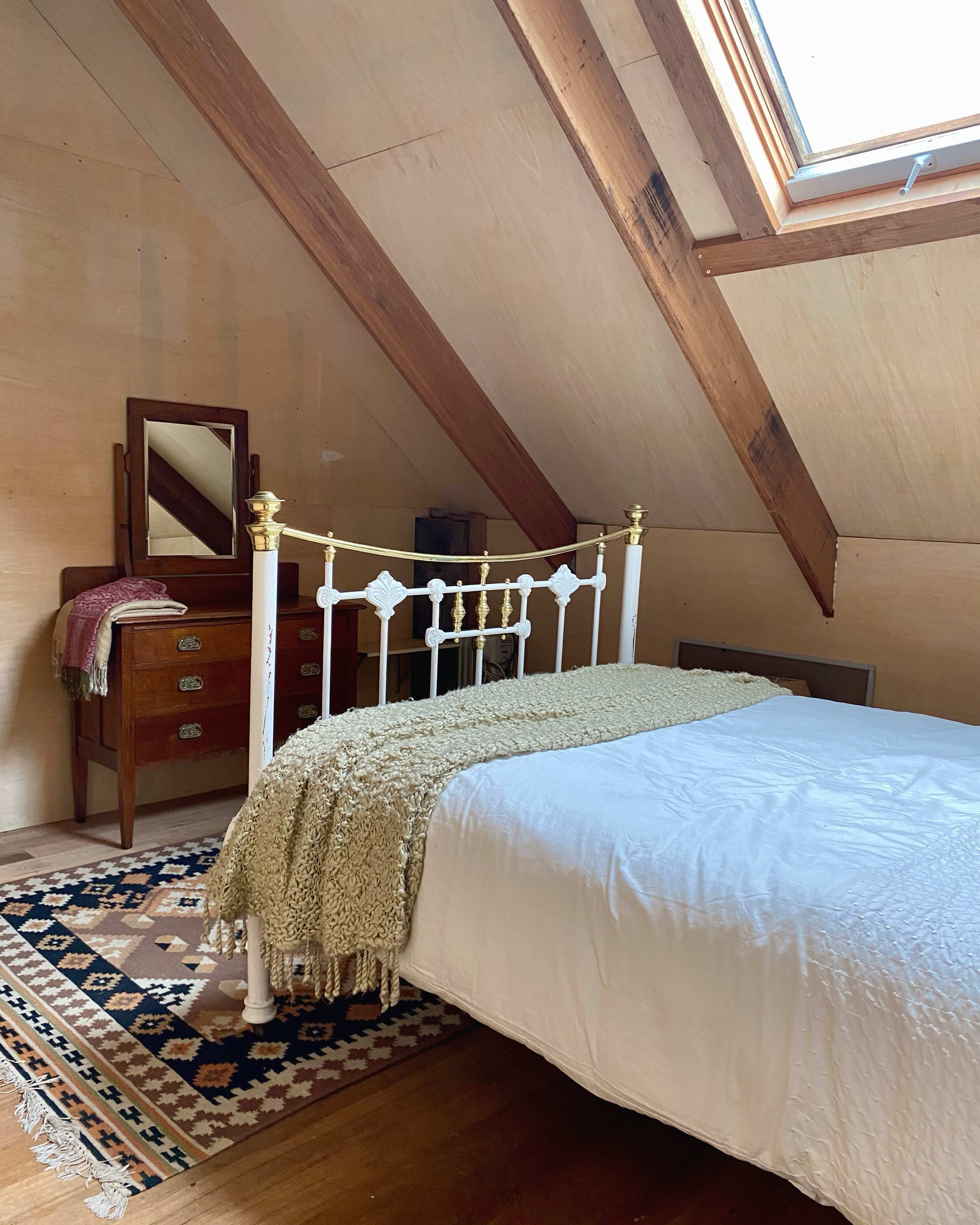 A cozy attic bedroom featuring a white metal bed with a patterned blanket, a wooden dresser, and a mirror under a slanted roof with exposed beams.