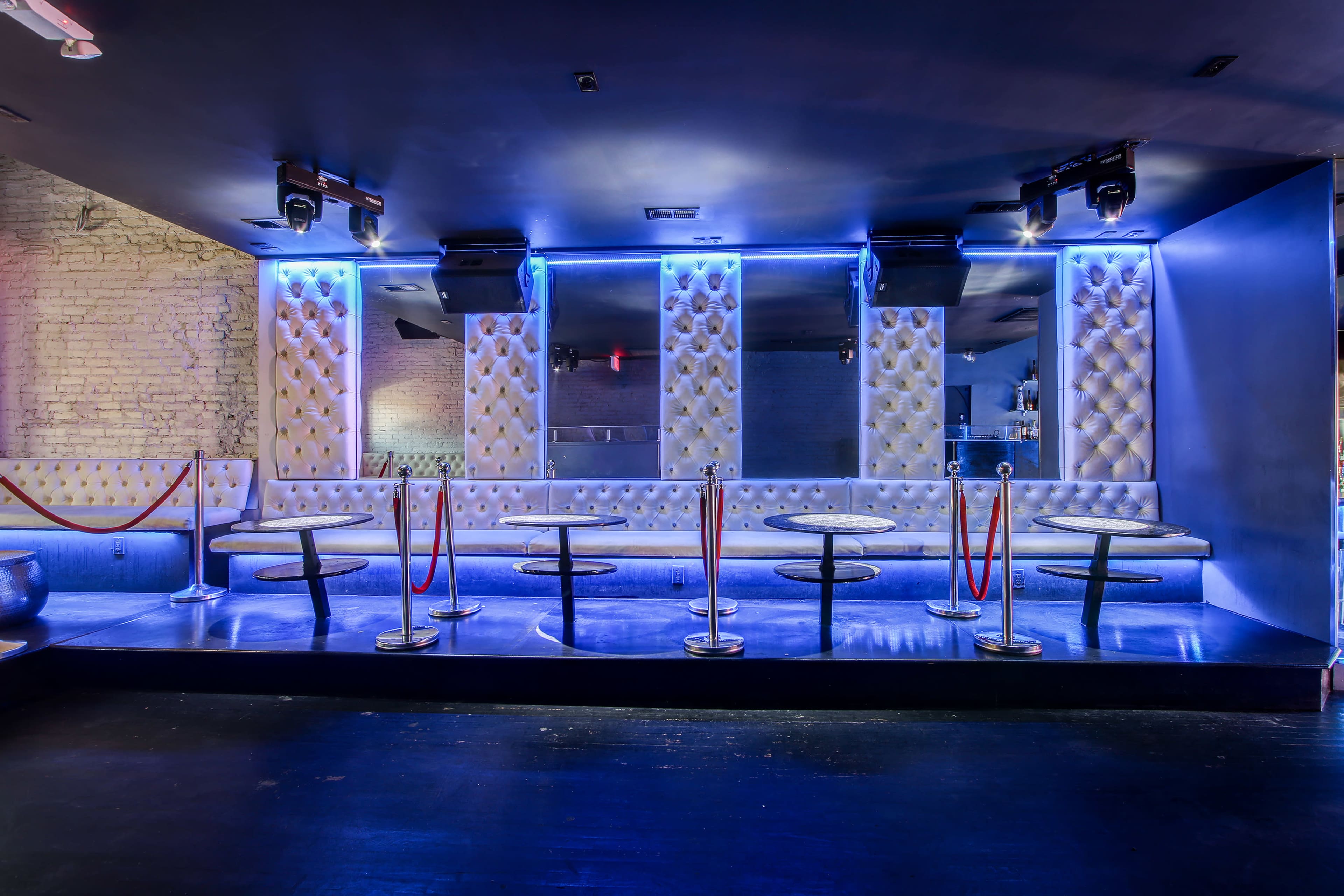 A dimly lit nightclub with padded walls, circular tables, and red ropes separating the seating area.