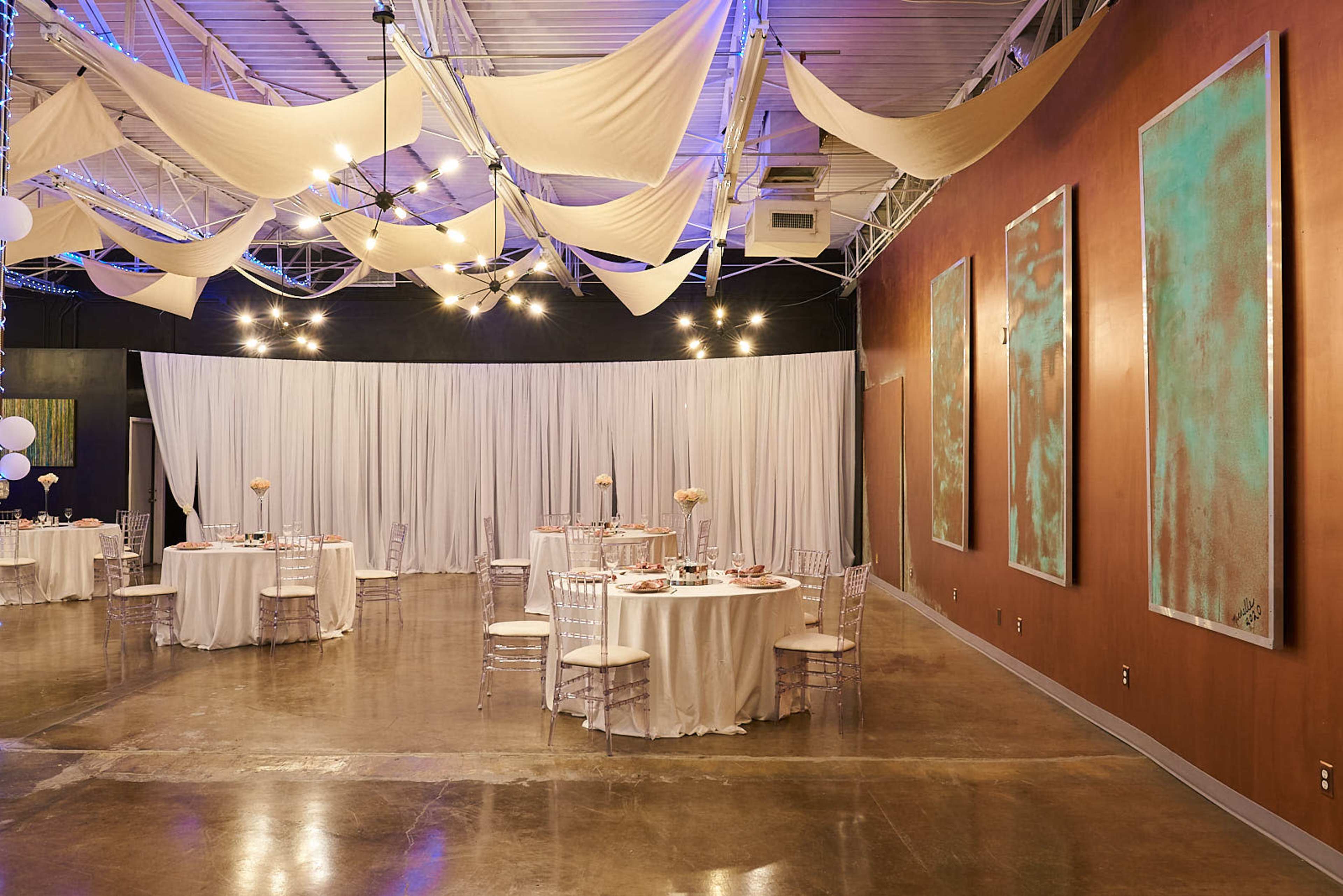 An indoor event space with round dining tables covered in white tablecloths, chair covers, and decorative centerpieces, alongside a backdrop of white drapery and modern artwork on the walls.