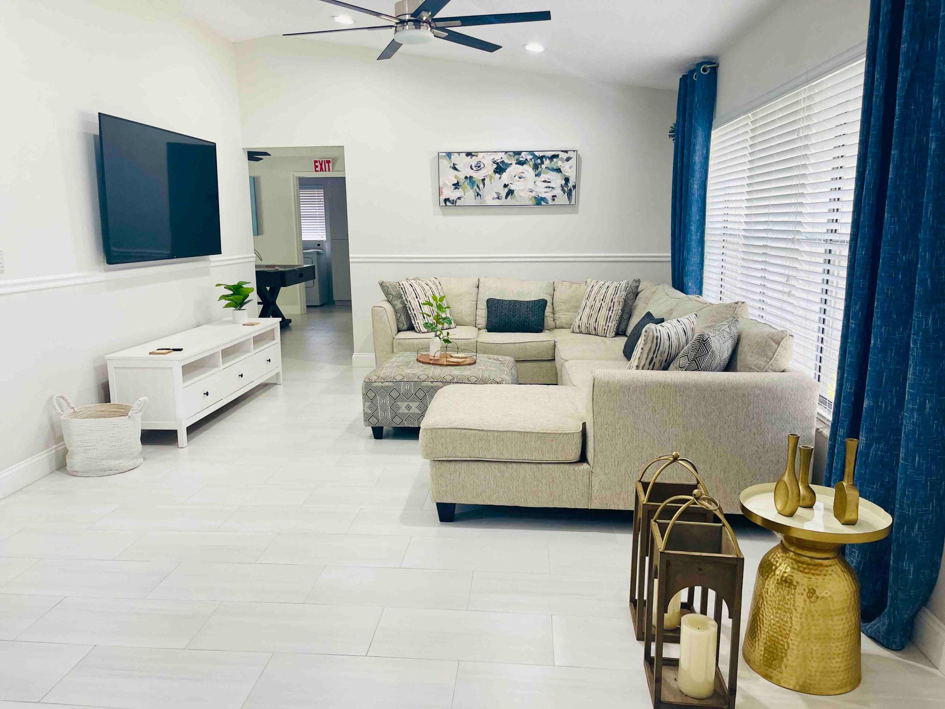 The image shows a modern living room with a sectional couch, a television mounted on the wall, and decorative furnishings including a gold side table and a plant.
