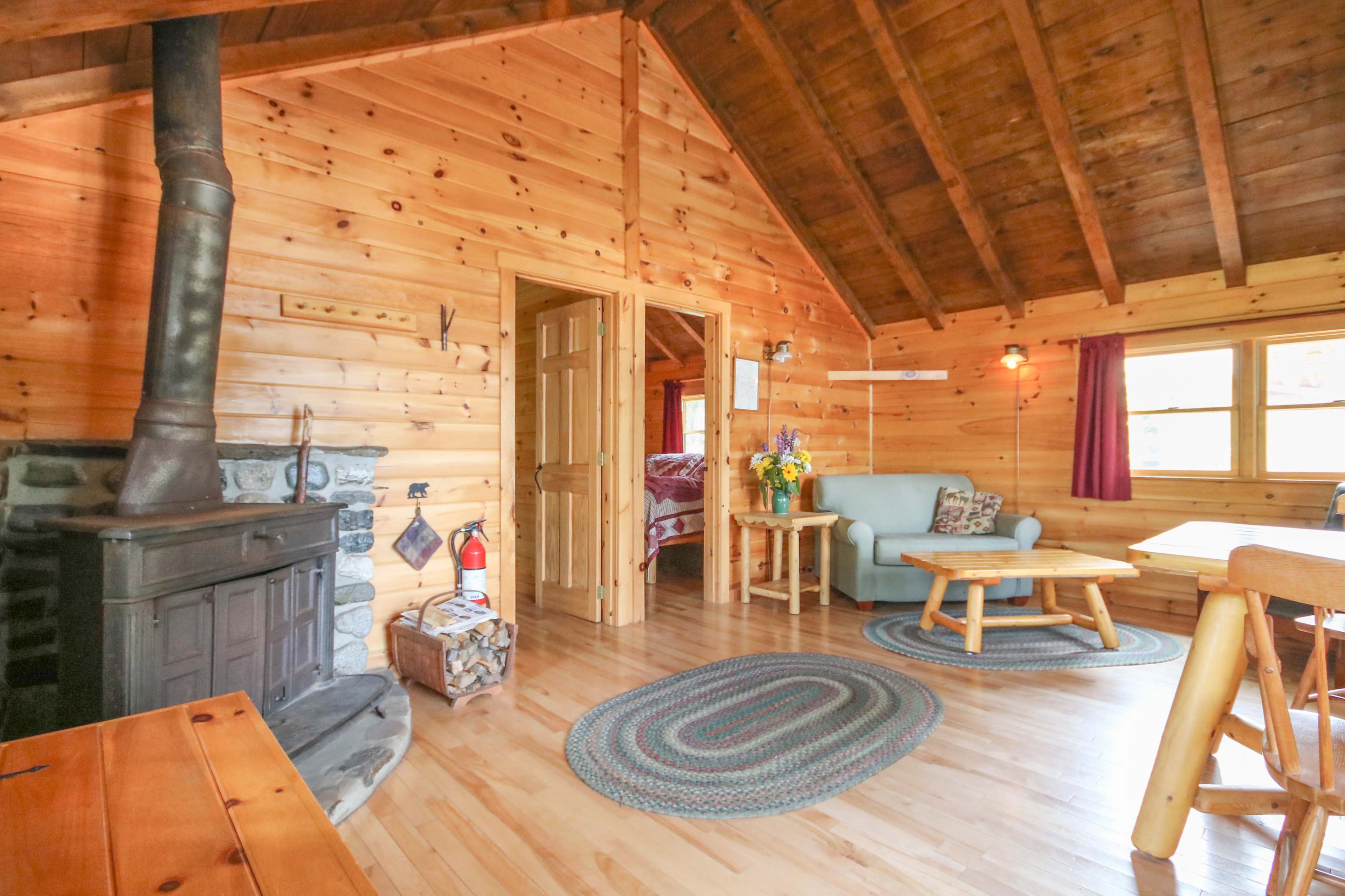 The interior of a wooden cabin features a living area with a sofa, a small table, and a wood stove, alongside a doorway leading to a bedroom.