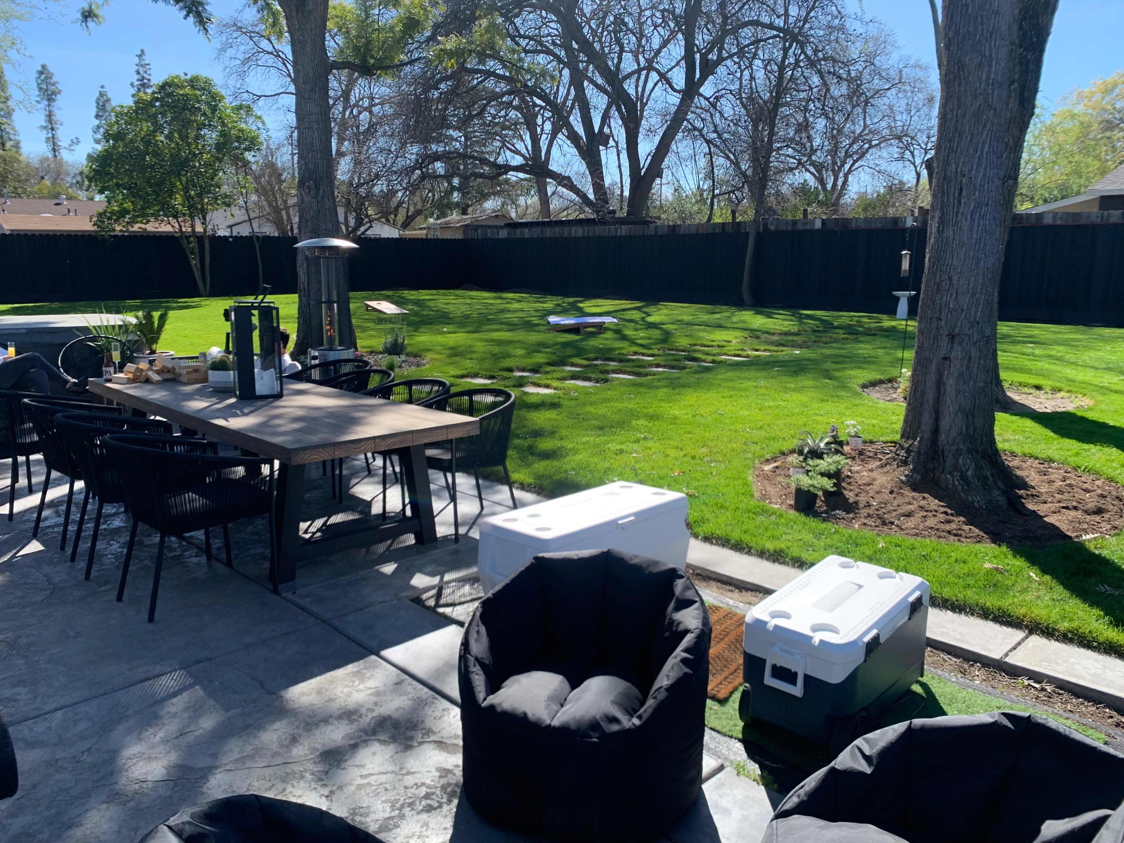 A patio area features a large table with chairs, coolers, and bean bag chairs, leading to a grassy backyard enclosed by trees and a black fence.
