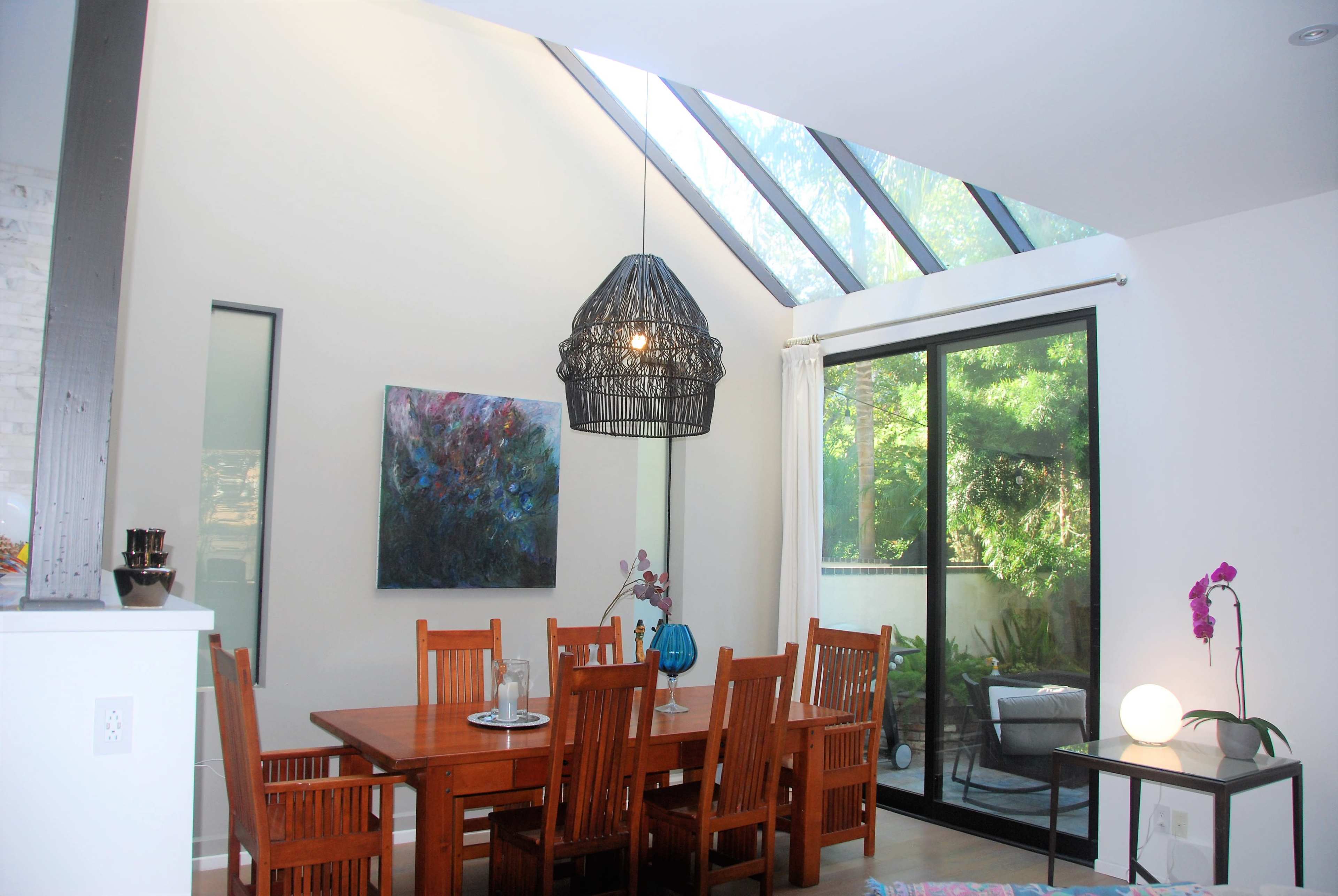 The image shows a modern dining area with a wooden table and chairs, a large window letting in natural light, and a hanging pendant light above the table.