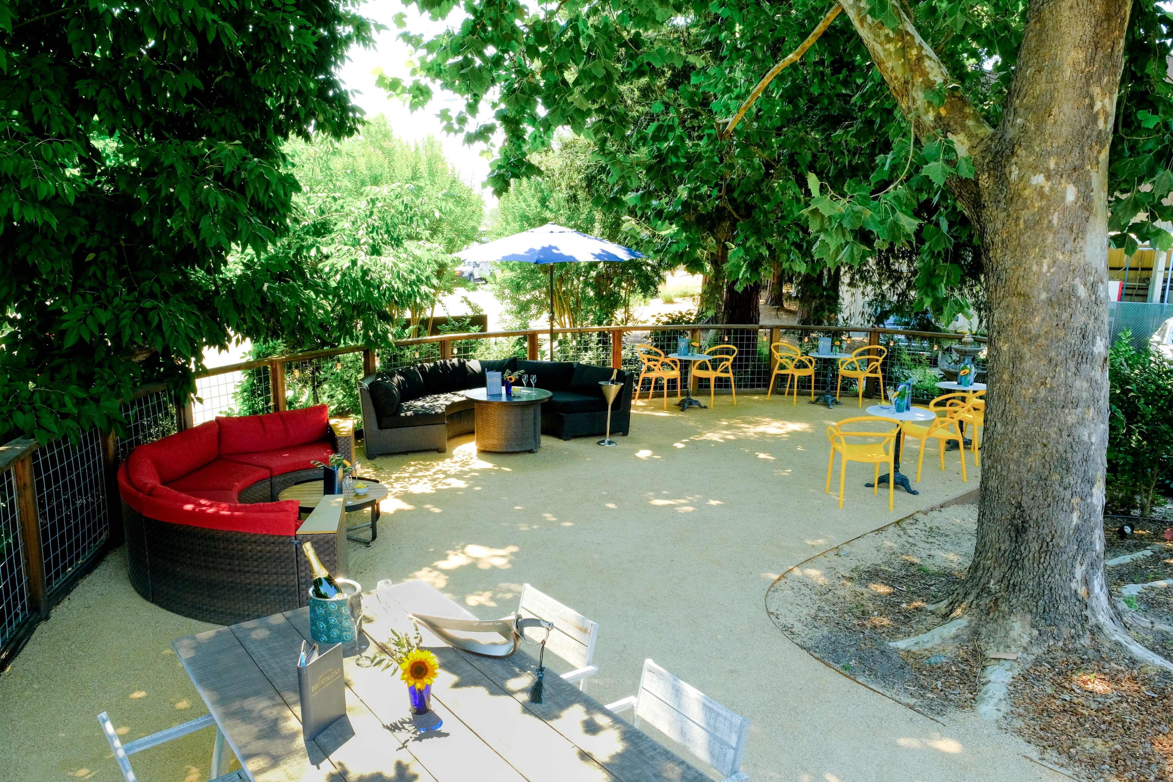 The image shows an outdoor seating area with various tables and chairs, including a red couch and a blue umbrella, surrounded by trees.