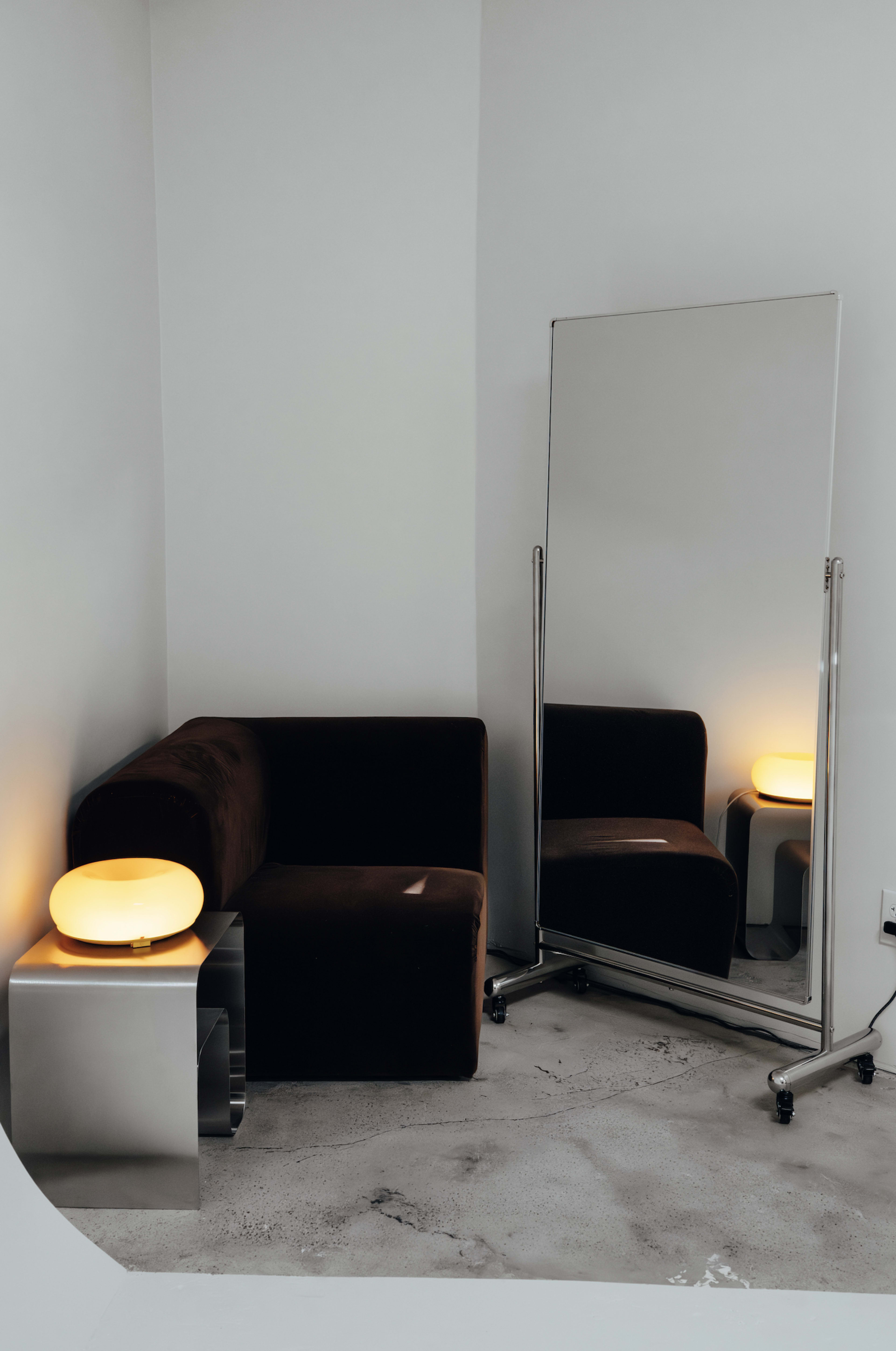 A dark velvet sofa and a large mirror are positioned near a soft-glowing lamp on a sleek side table, set against a light-colored wall.